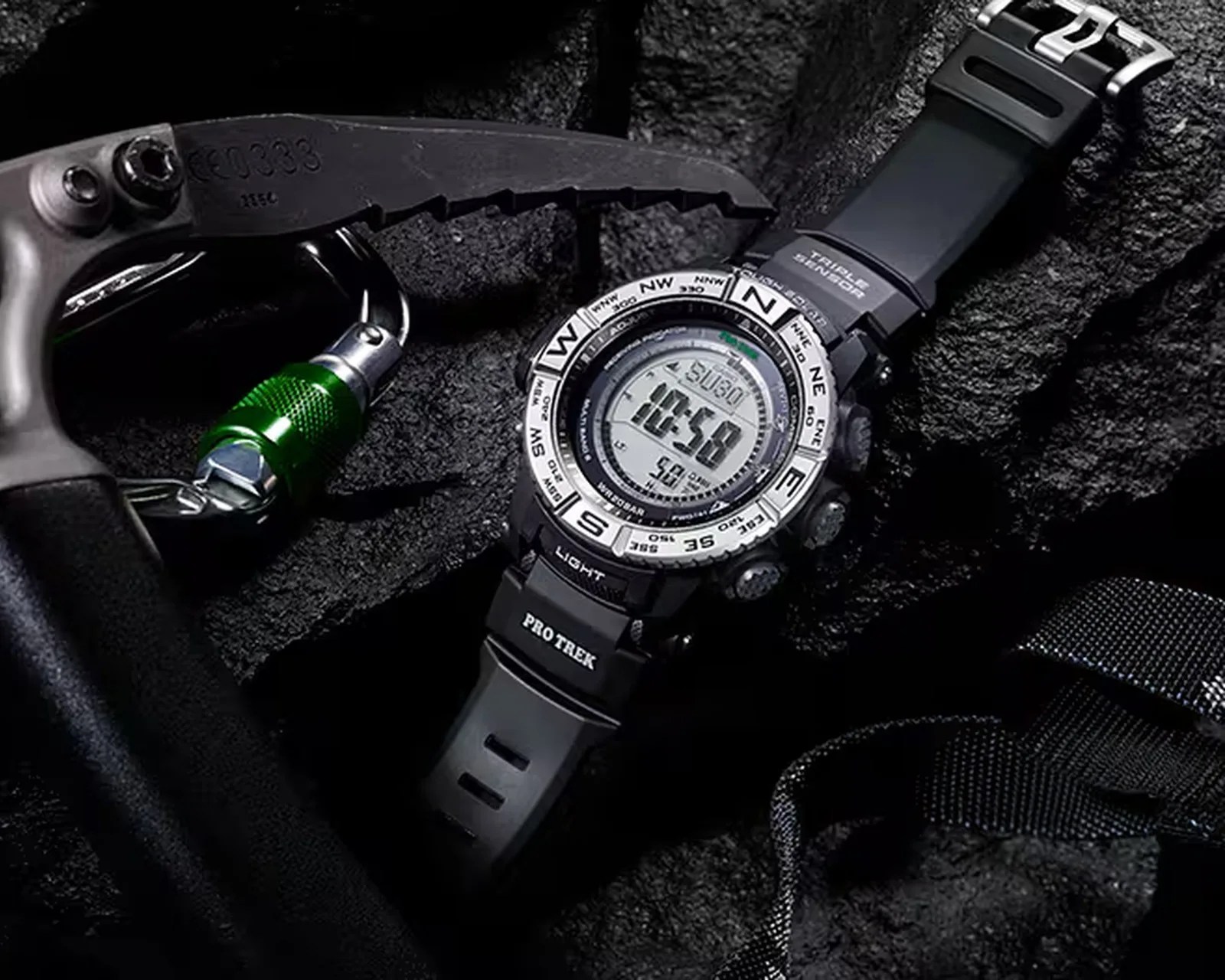 Black Casio Pro Trek digital watch with compass bezel and rubber strap on textured black surface next to a climbing carabiner.