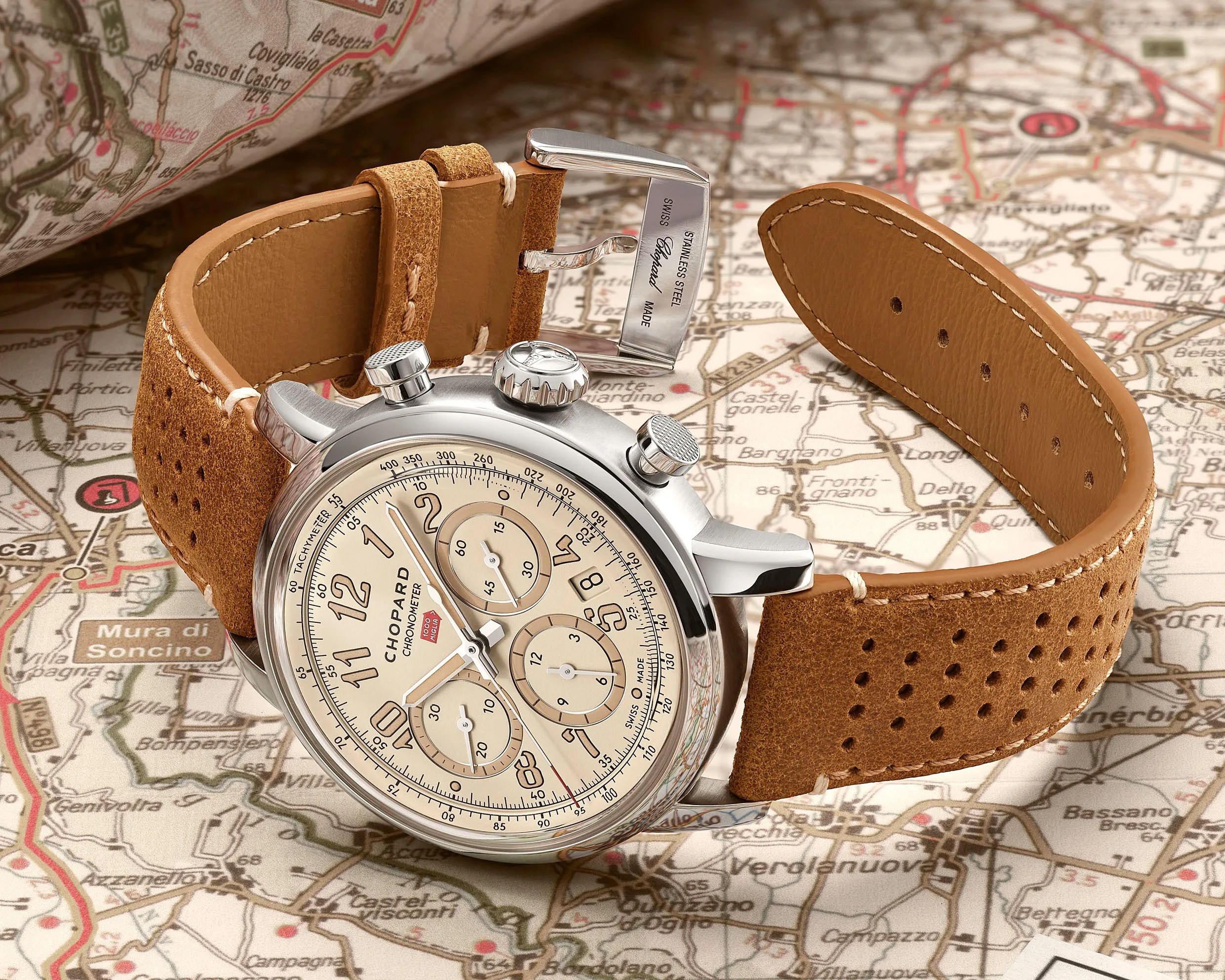 Chronograph wristwatch with beige dial, stainless steel case, and perforated tan leather strap on a map background.