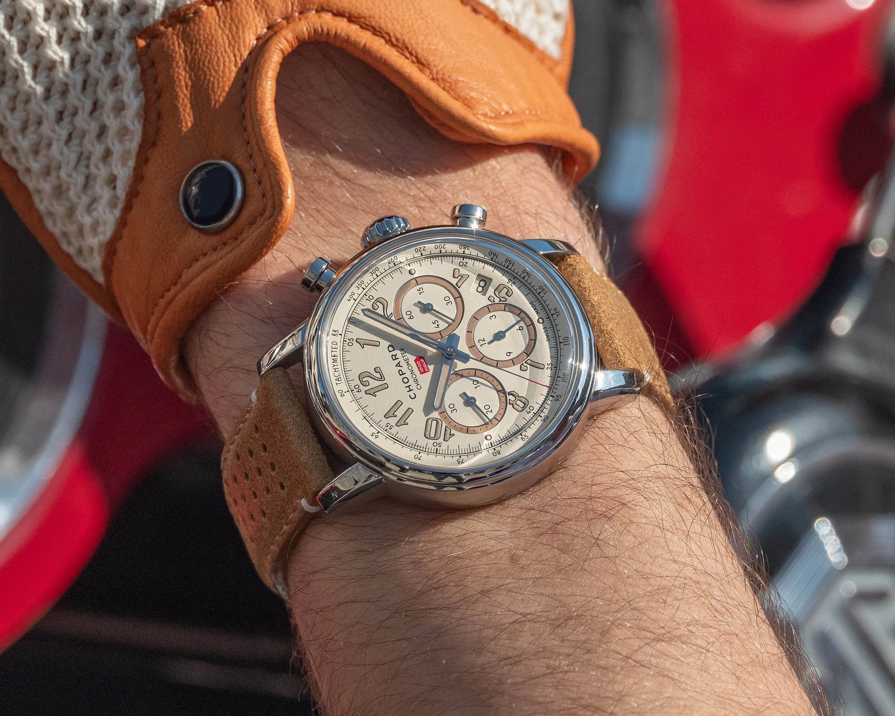 Silver Chopard chronometer wristwatch with beige dial and brown leather strap worn on a wrist with a tan driving glove.
