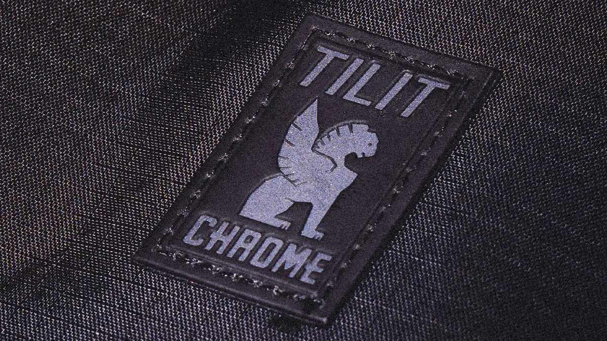 Black fabric with a stitched rectangular patch featuring a winged lion logo and the text "TILIT CHROME.