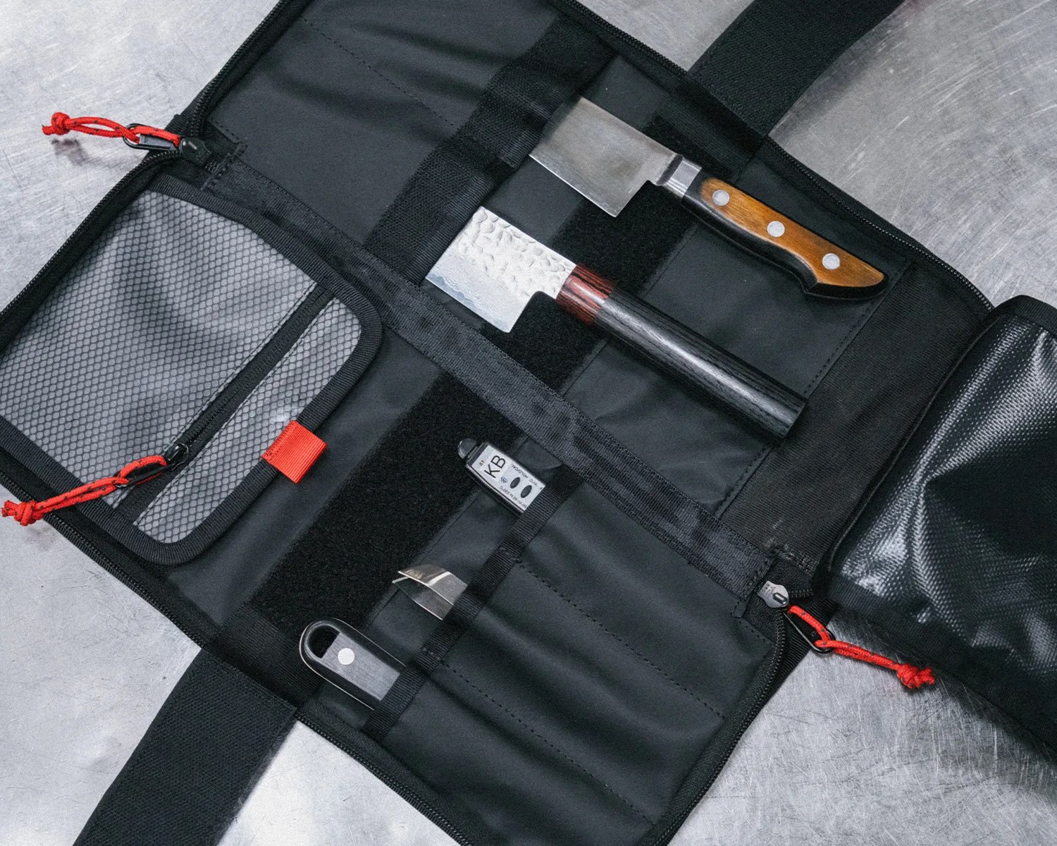 Black knife roll with red zipper pulls containing two chef knives and a small tool on a metal surface.