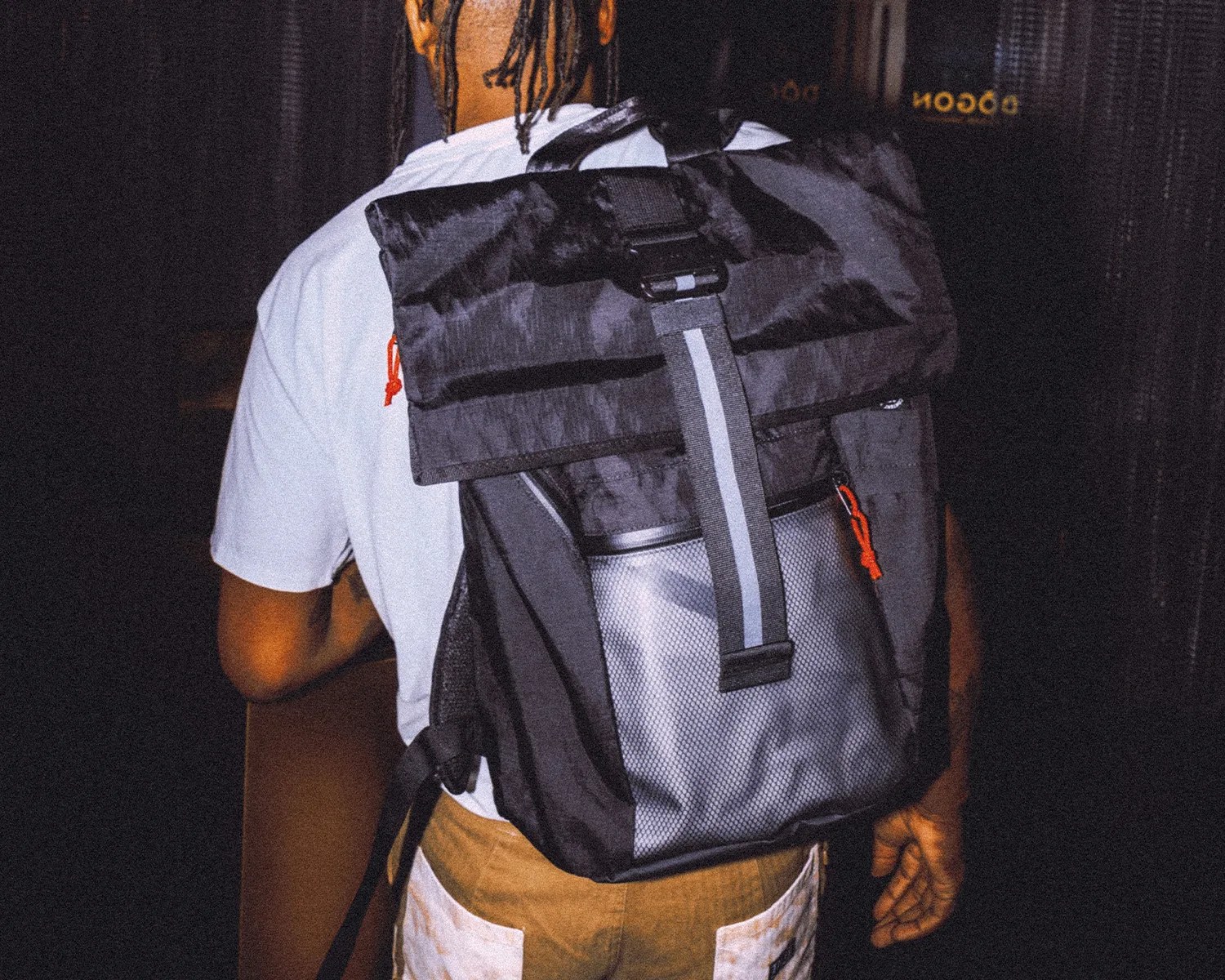 Black and gray roll-top backpack with reflective strap and orange zipper pulls worn by a person in a white shirt.