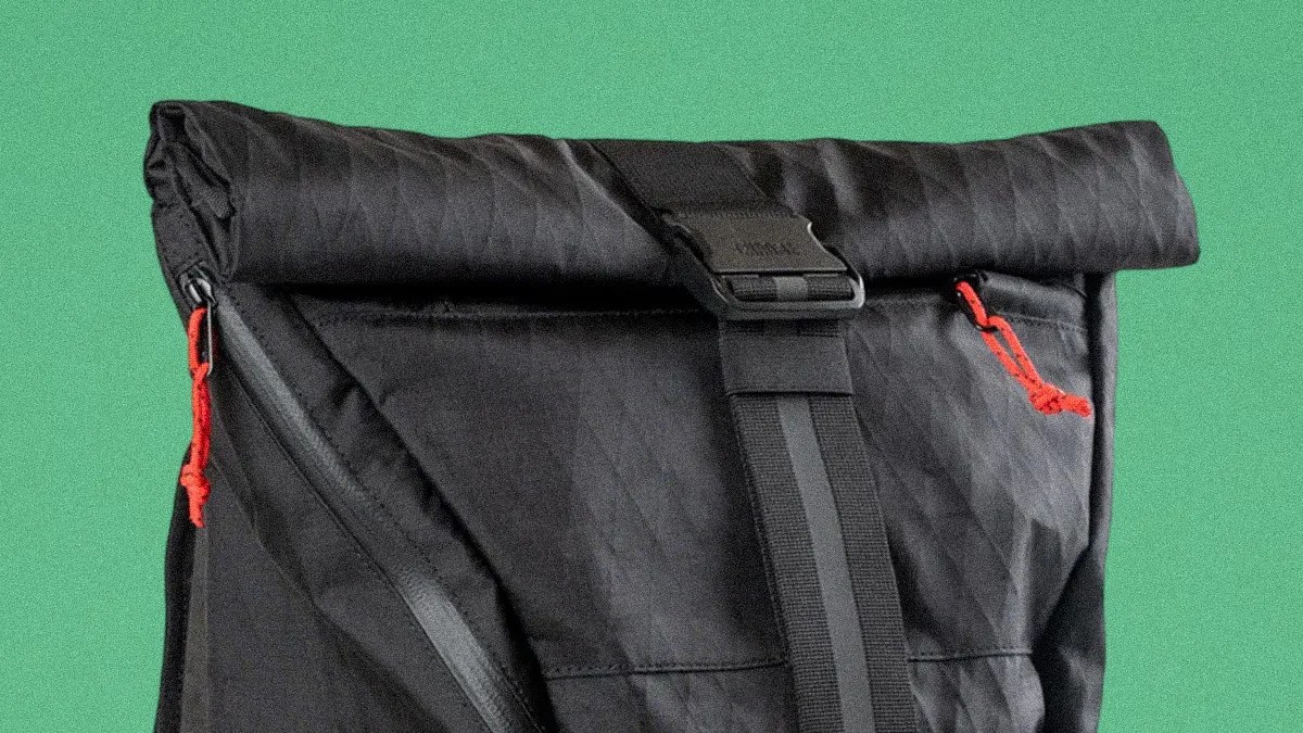 Black roll-top backpack with diagonal zippers and red zipper pulls against a green background.