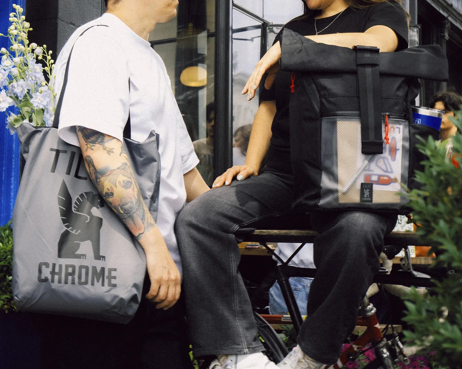 Two people with black and gray tote bags, one with flowers inside, sitting and standing near a cafe window.