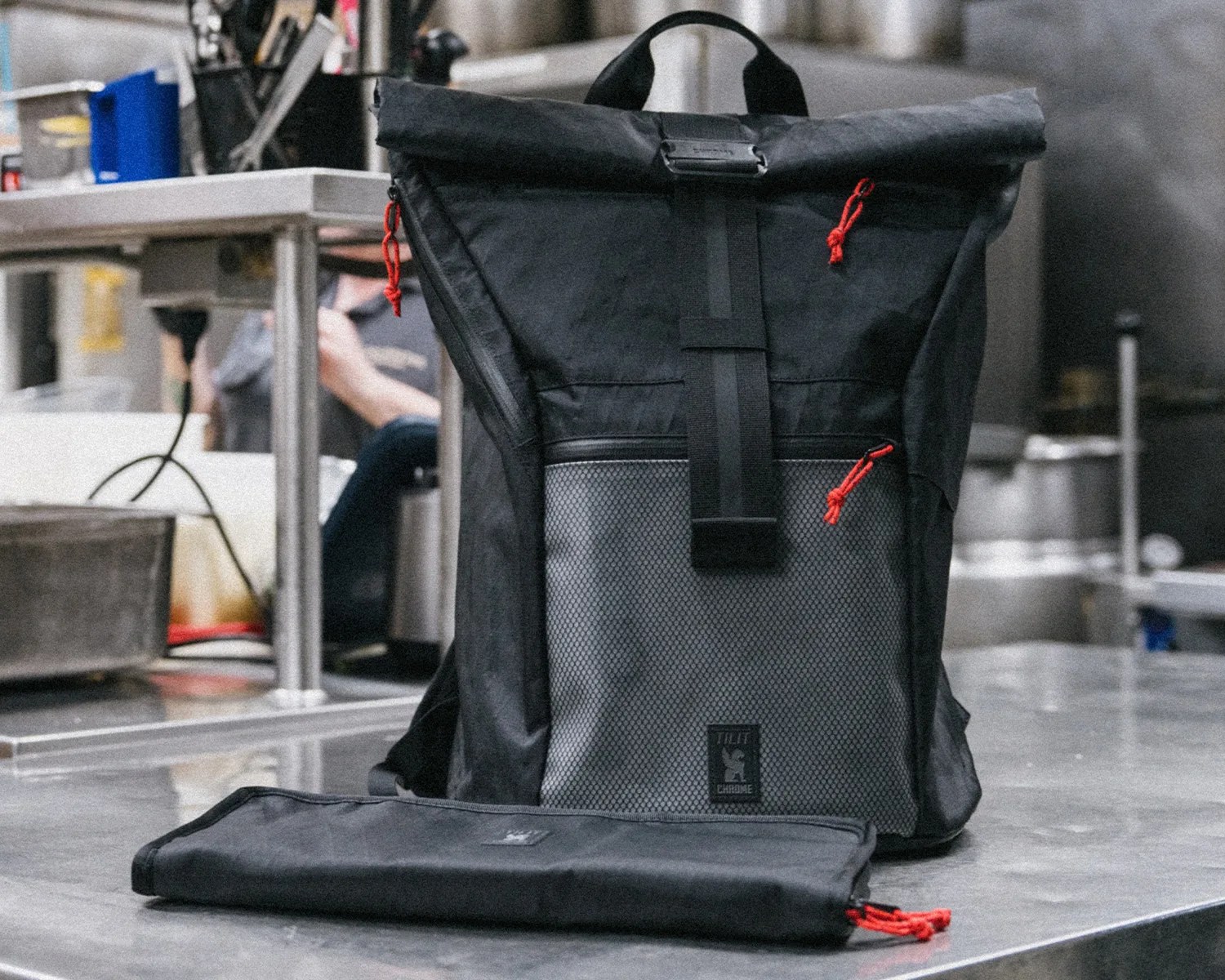 Black roll-top backpack with gray front pocket and red zipper pulls on a metal surface.