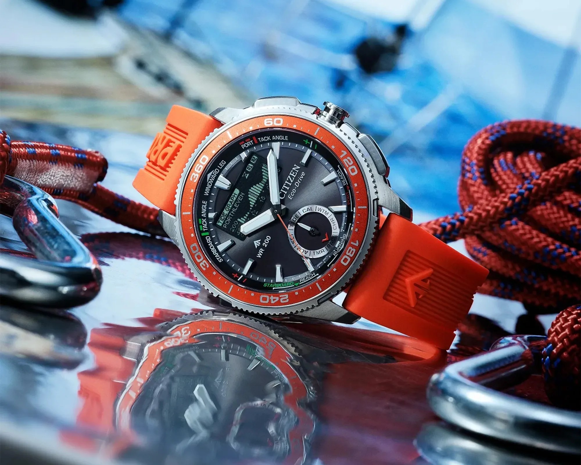Close-up of a Citizen Eco-Drive wristwatch with an orange bezel and strap, resting on a reflective surface near red and blue ropes.