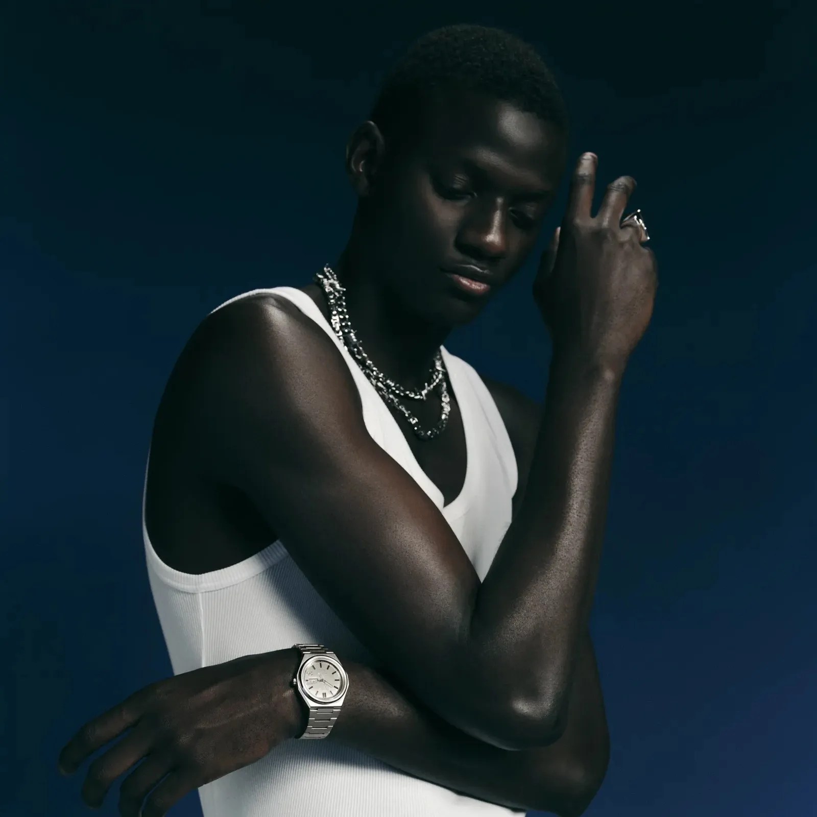 Person wearing a white sleeveless top with silver chain necklaces, a silver watch, and a silver ring against a dark blue background.