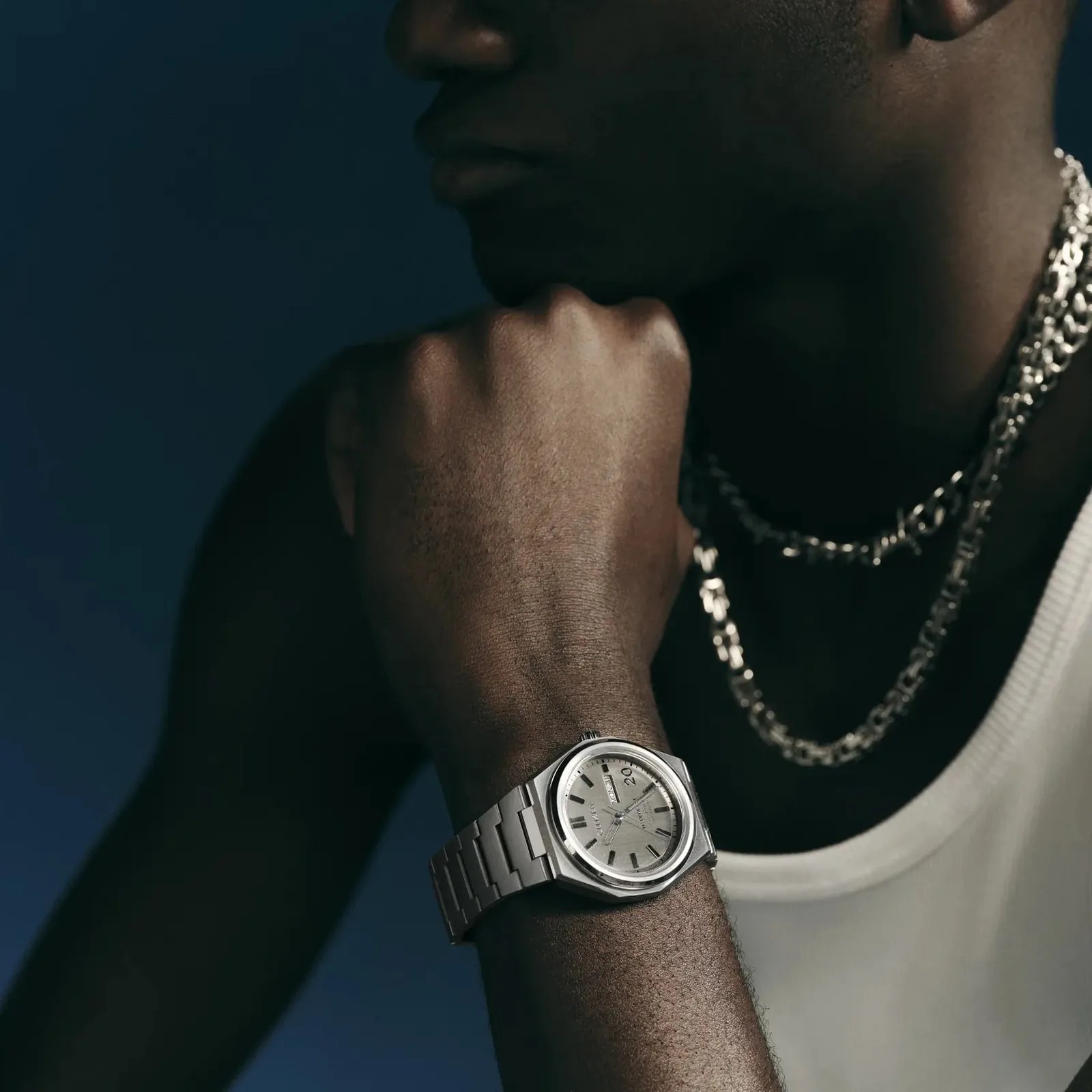 Close-up of a wrist wearing a silver Citizen watch with a metal band and a silver chain necklace on a person in a white tank top.