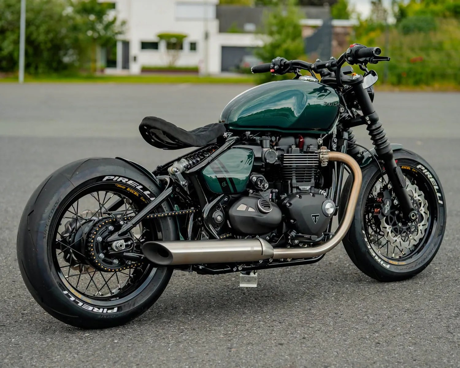 Dark green custom cafe racer motorcycle with black frame, Pirelli tires, and silver exhaust pipe on asphalt.