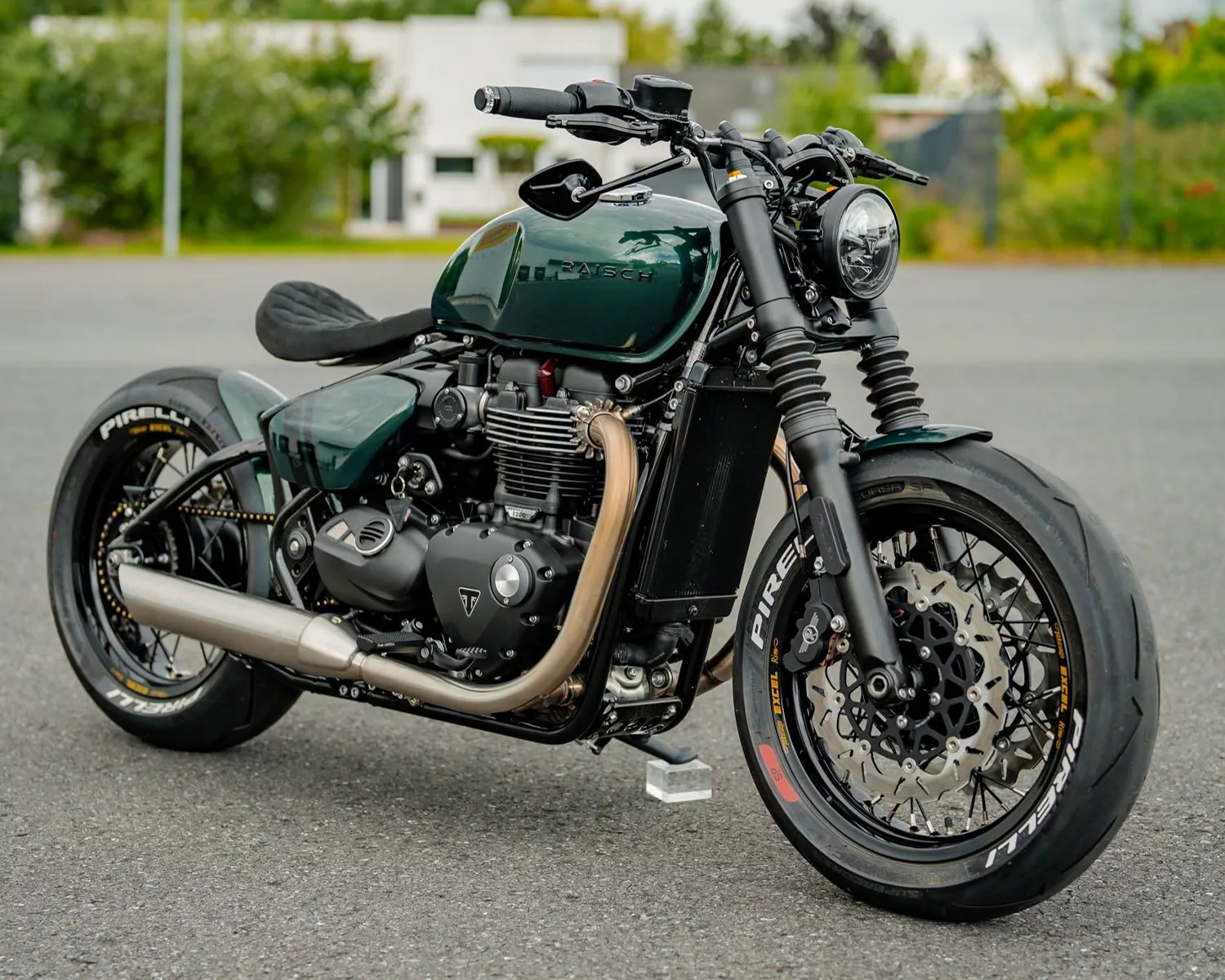 Dark green custom Triumph motorcycle with black frame, Pirelli tires, and exposed engine on asphalt.