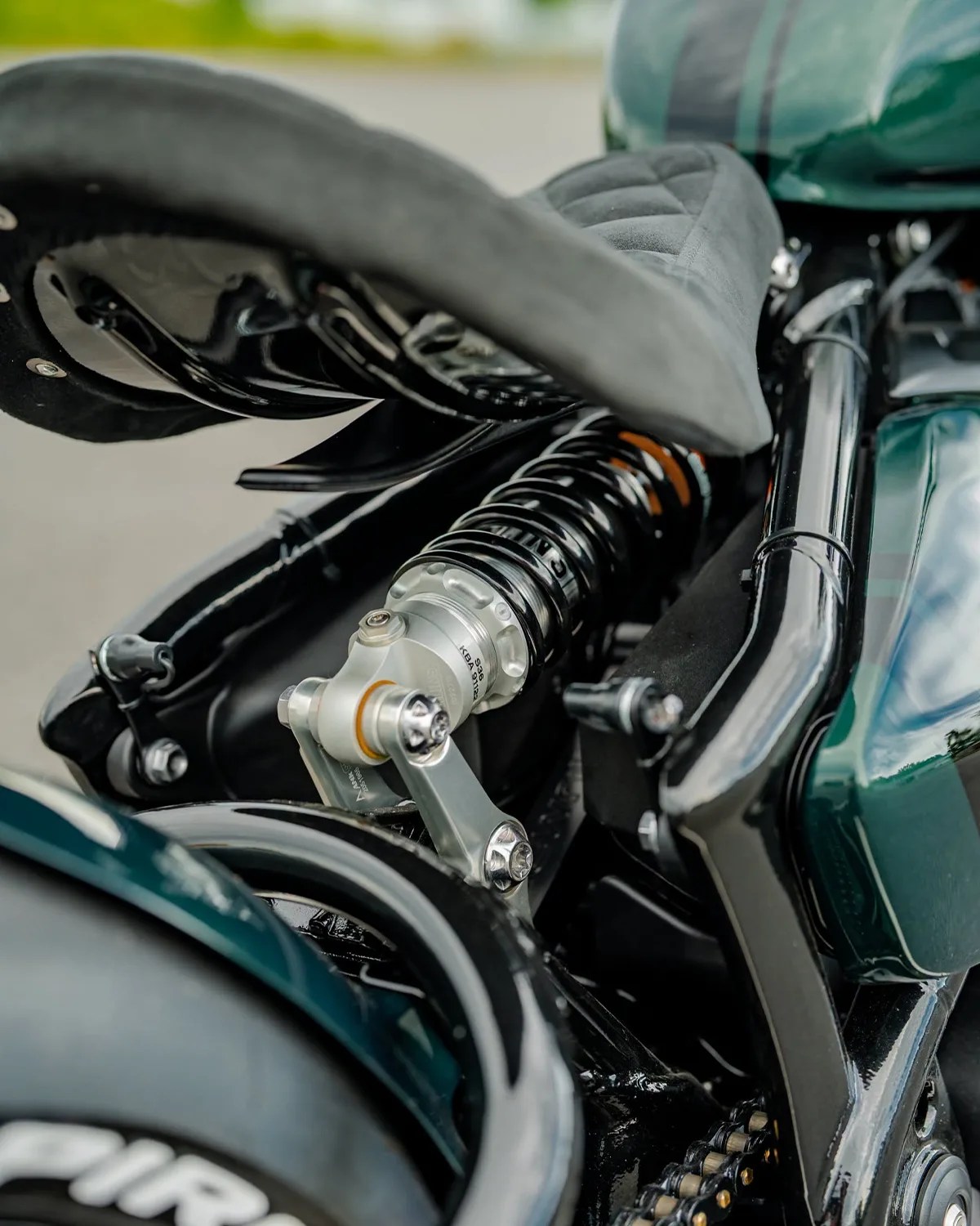 Close-up of a black coil spring shock absorber mounted under a motorcycle seat with visible chain and green body parts.
