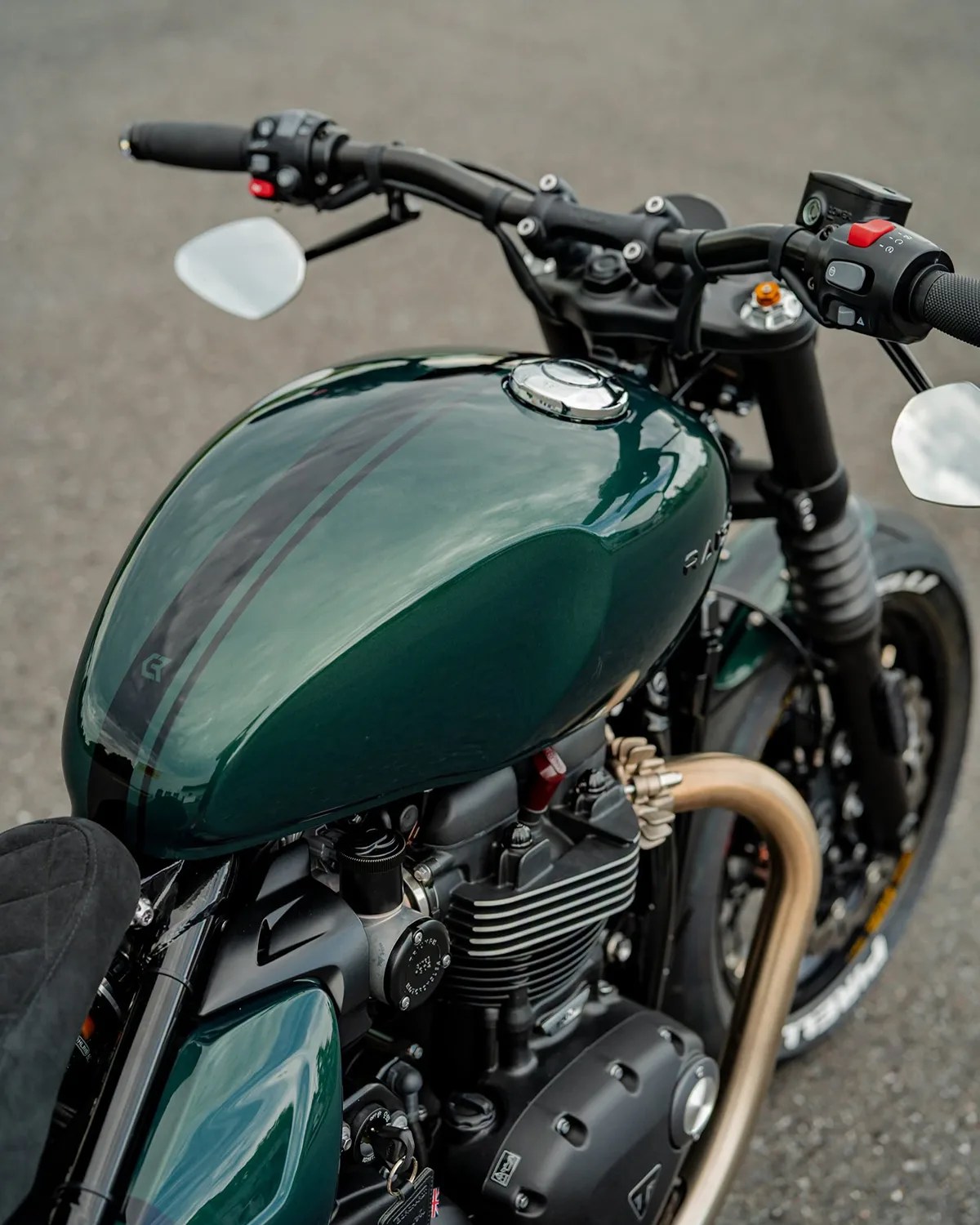 Glossy dark green motorcycle fuel tank with black racing stripes and black handlebars on a gray asphalt surface.