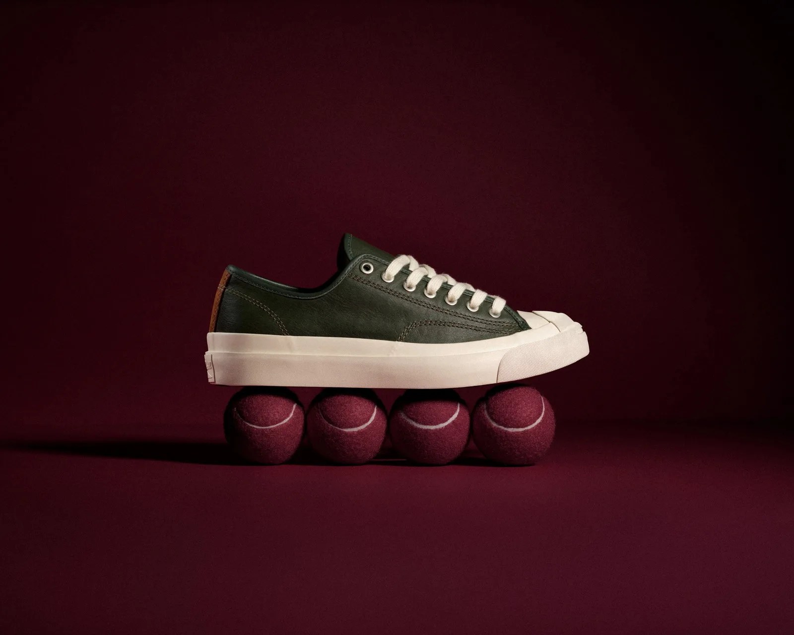 Dark green low-top sneaker with white laces and sole balanced on four maroon tennis balls.