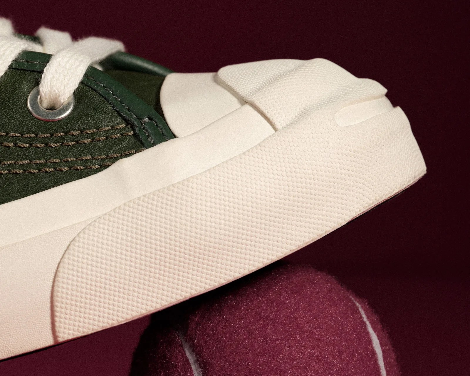 Close-up of a green sneaker with white laces and a textured off-white sole resting on a maroon tennis ball.