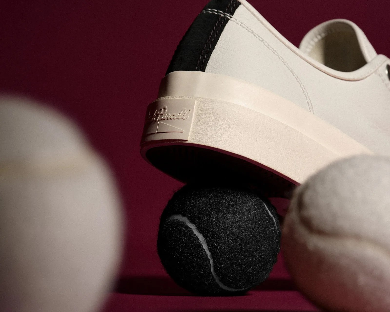 Off-white sneaker with black heel detail balanced on a black tennis ball against a dark red background.