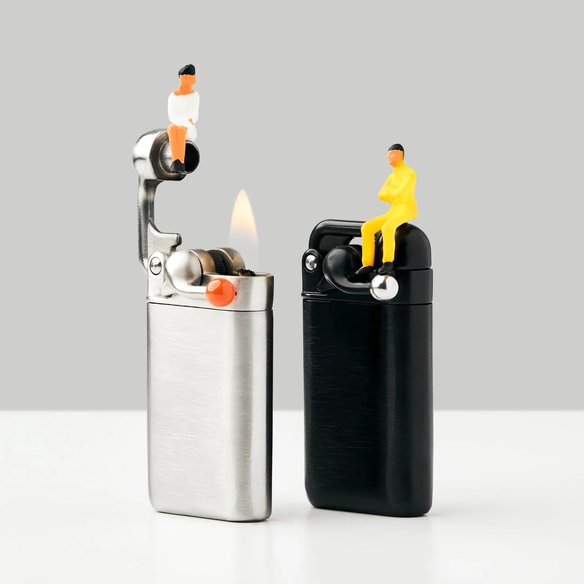 Silver and black lighters with miniature figurines sitting on top against a gray background.