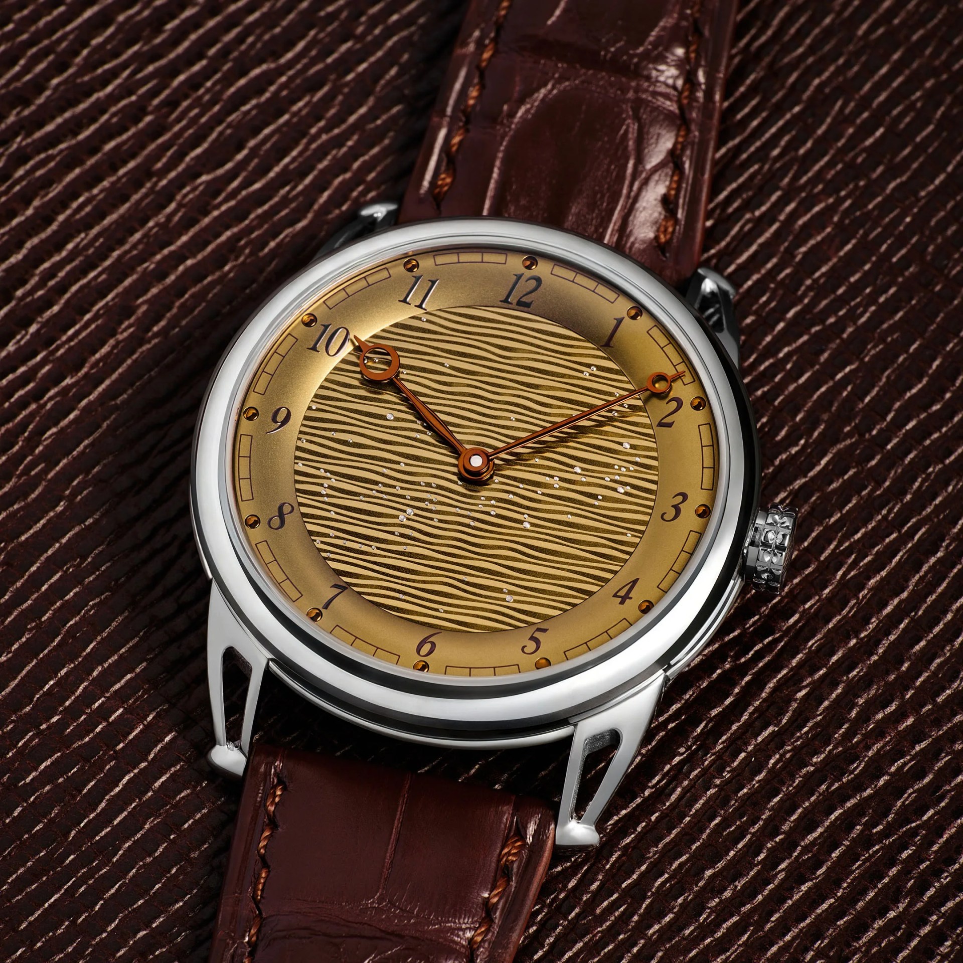 Round silver watch with gold textured dial and brown leather strap on a brown textured surface.