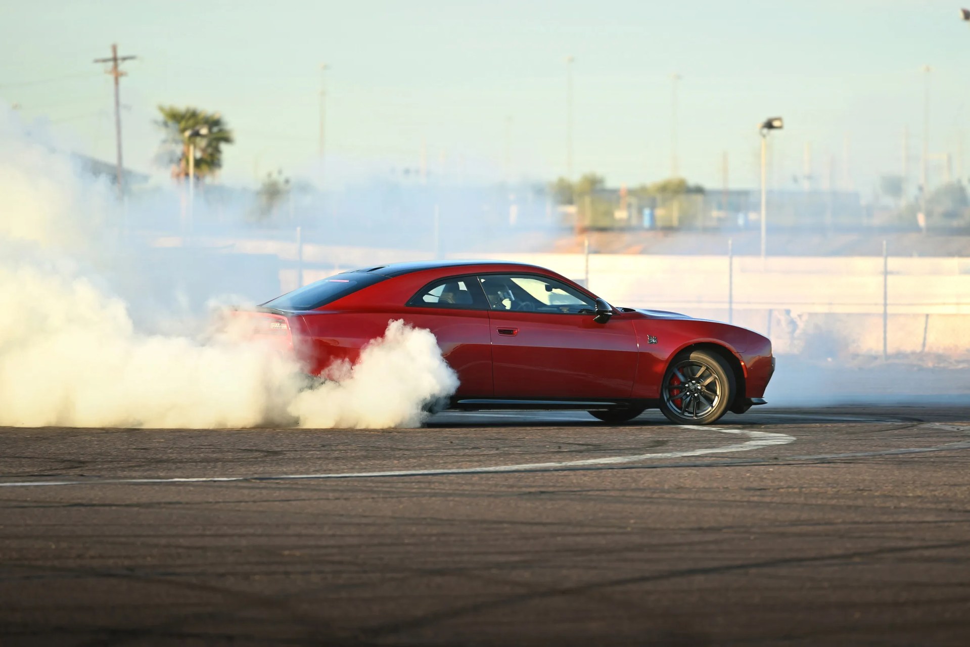 Red Dodge Challenger muscle car performing a burnout with smoke around the rear tires on a paved surface.