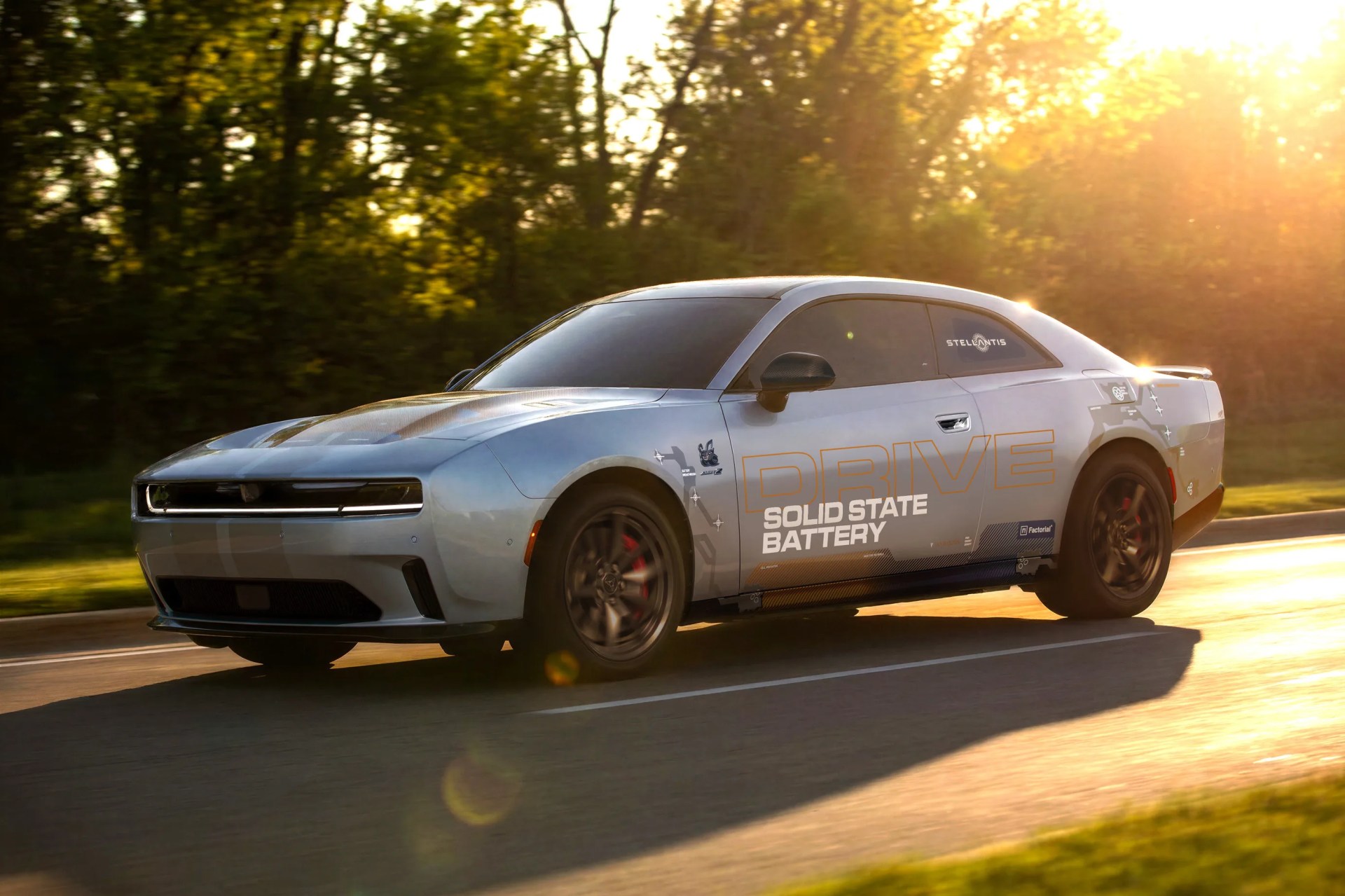 Silver electric muscle car with "DRIVE SOLID STATE BATTERY" text on the side driving on a sunlit road.