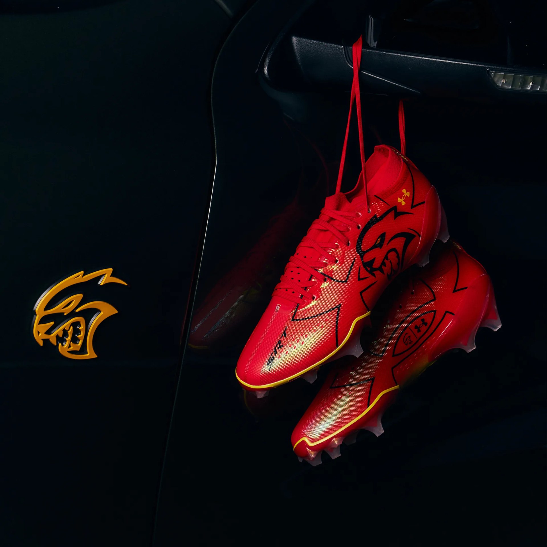Red Under Armour cleats with black and yellow accents hanging by their laces against a dark background with a yellow stylized cat logo.