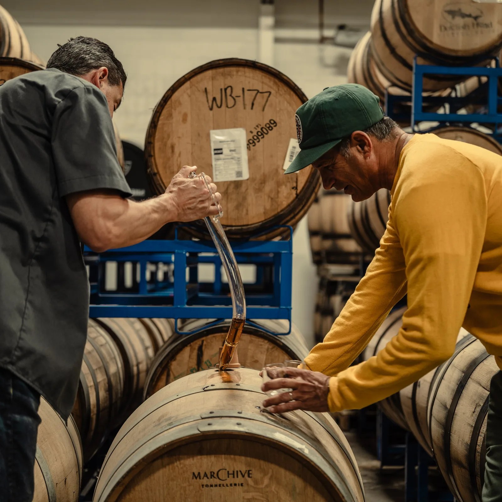 Two men inspecting liquid being drawn from a wooden barrel in a warehouse filled with stacked barrels.