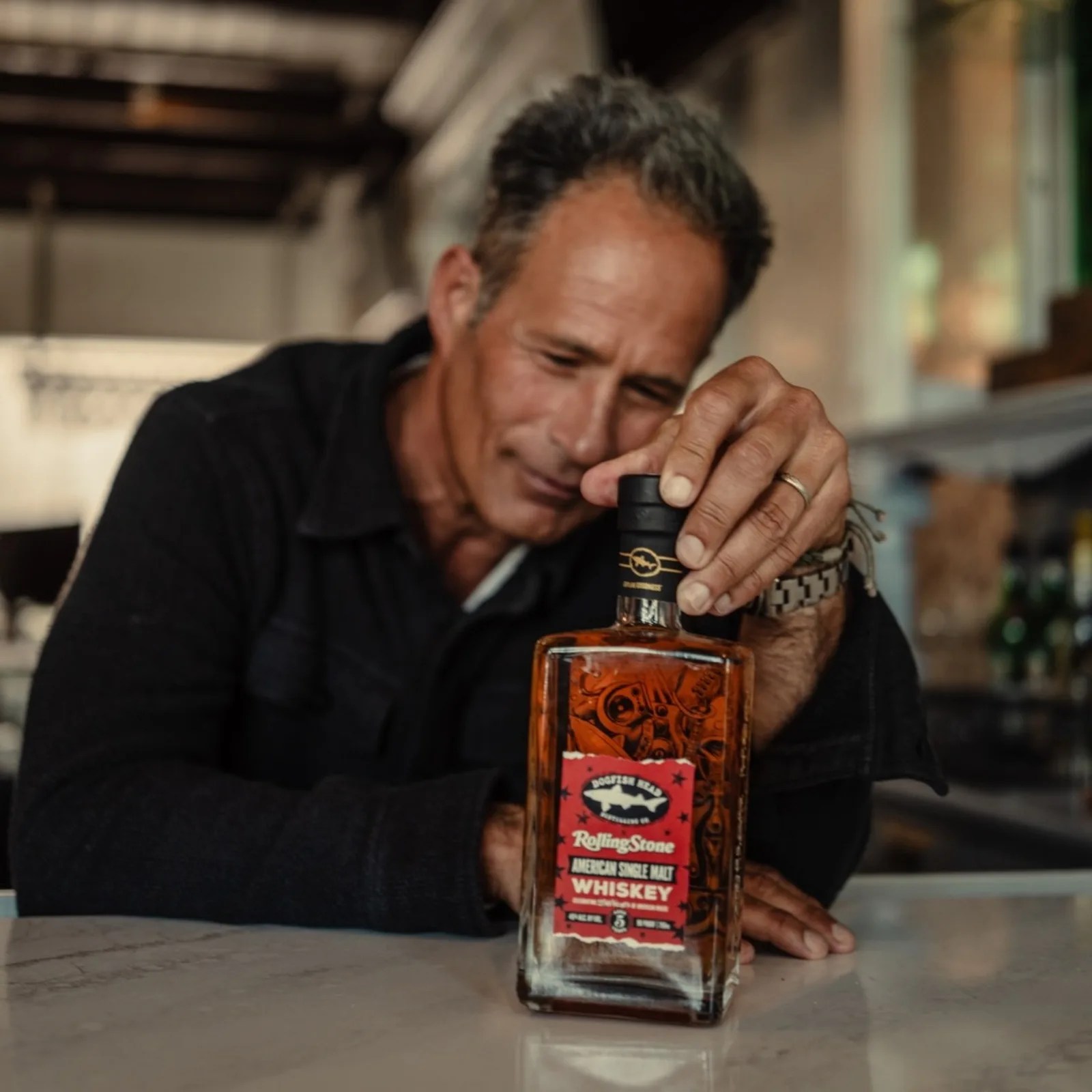Man in a black shirt holding a bottle of Dogfish Head Rolling Stone American Single Malt Whiskey on a marble surface.