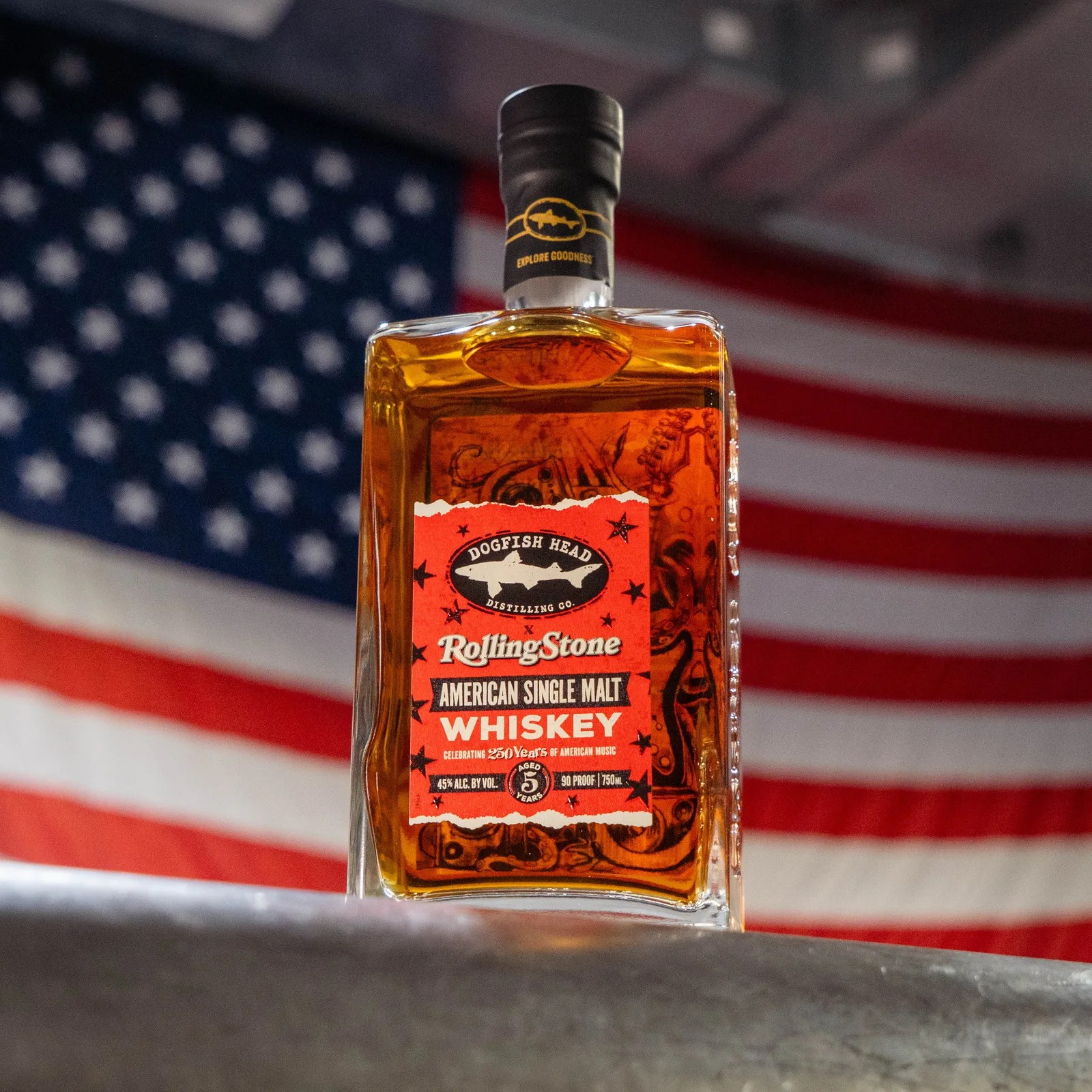 Rectangular bottle of Dogfish Head Rolling Stone American Single Malt Whiskey with red label in front of a blurred American flag.