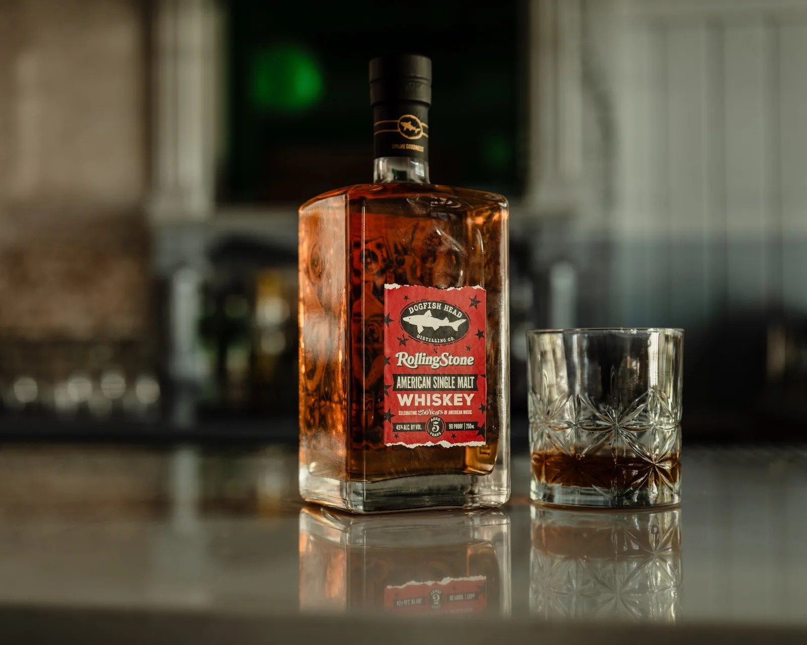 Square bottle of Dogfish Head Rolling Stone American Single Malt Whiskey next to a glass with whiskey on a reflective surface.