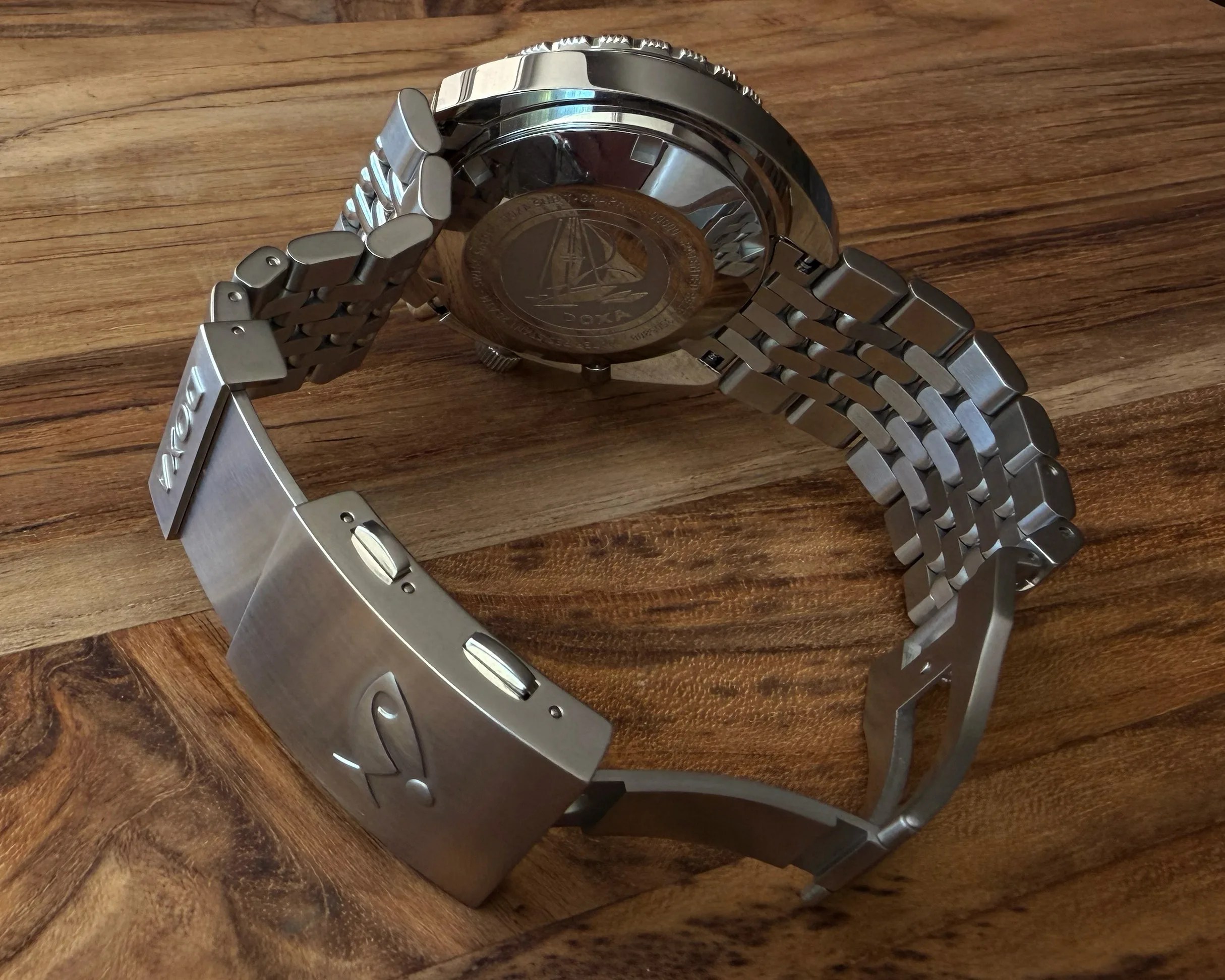 Stainless steel watch bracelet with a clasp engraved with "DOXA" on a wooden surface.