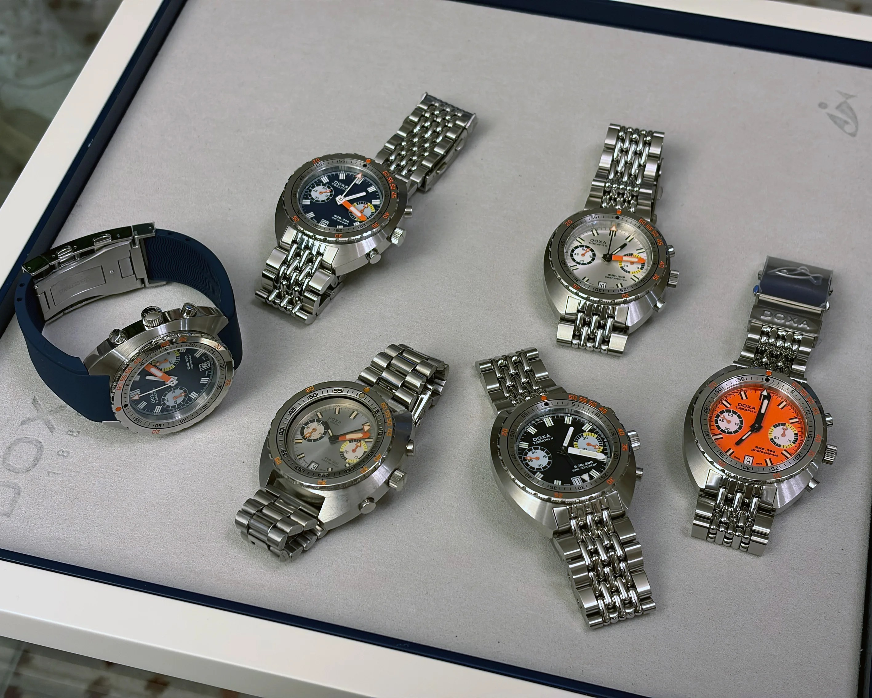 Seven stainless steel Doxa wristwatches with various dial colors and metal bracelets displayed on a light gray surface.