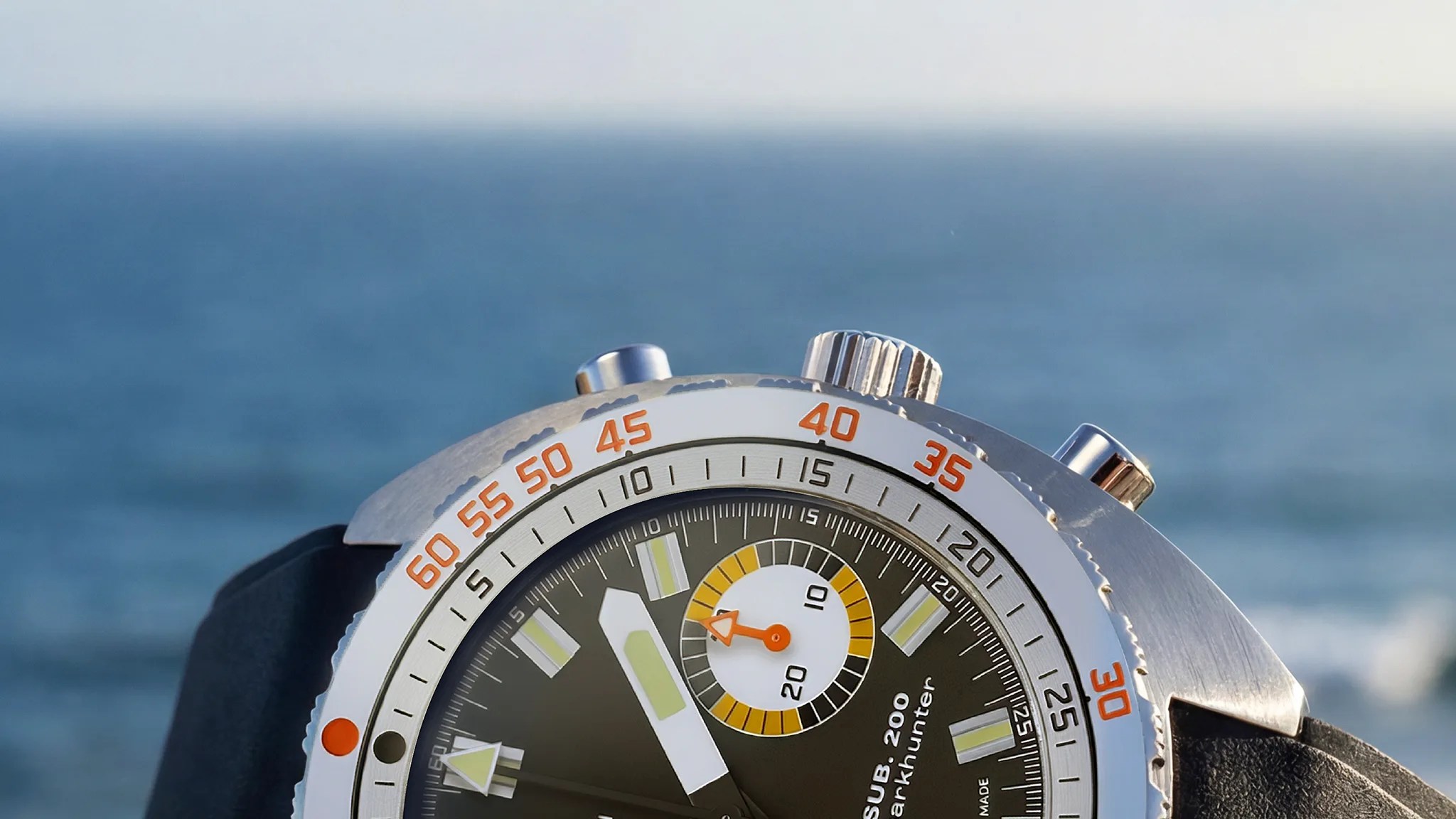 Close-up of a stainless steel dive watch bezel with orange minute markers and a black dial against a blue ocean background.