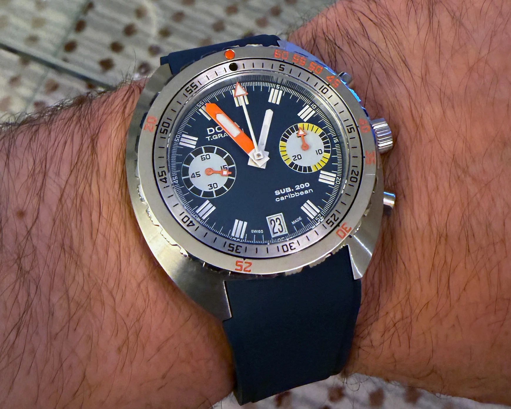Silver dive watch with black dial, orange and white hands, two subdials, and black strap on a wrist.