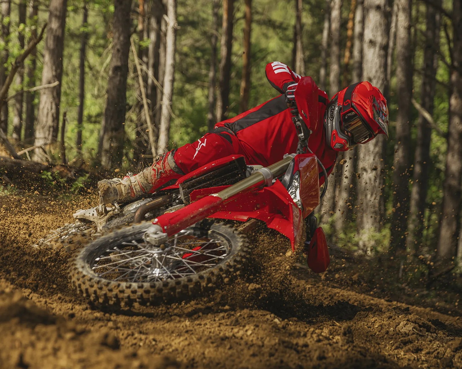 Dirt bike rider in red gear leaning sharply on a red motocross bike kicking up dirt in a forest.