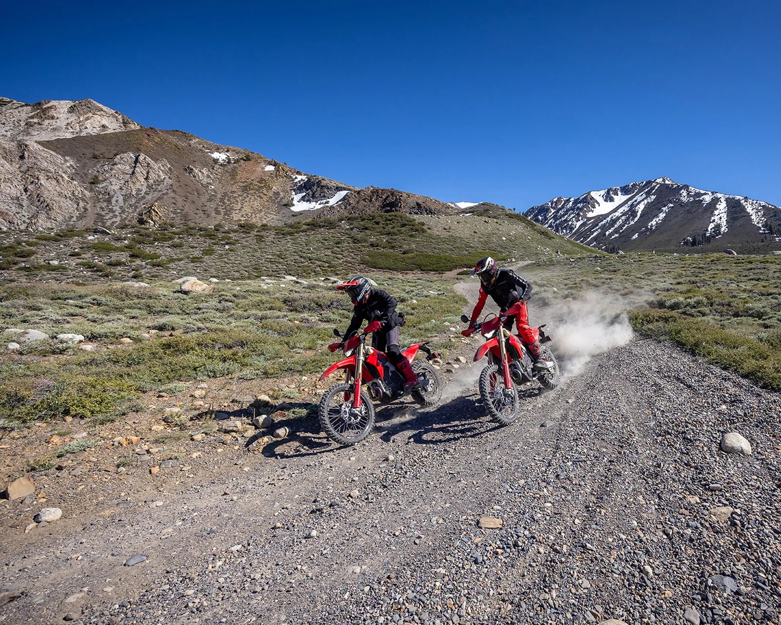 Two riders on red dirt bikes kicking up dust on a rocky trail with mountains in the background.
