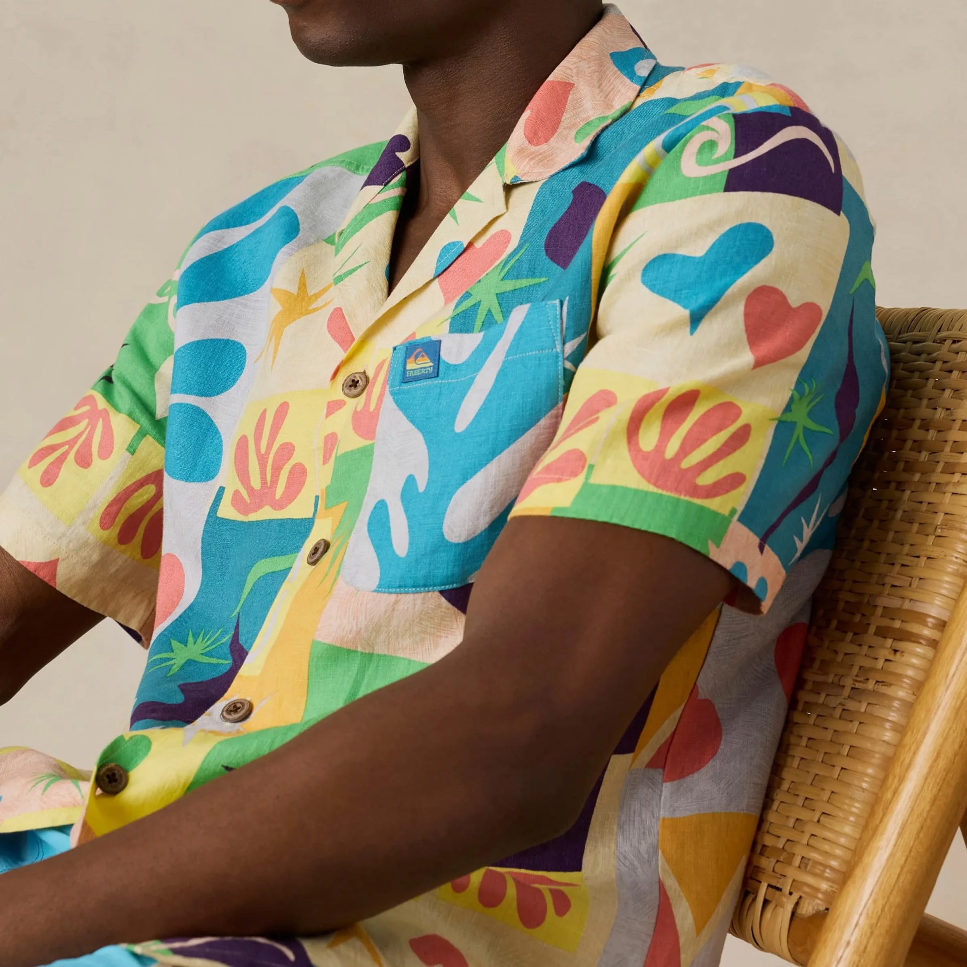 Short-sleeve button-up shirt with colorful abstract shapes and patterns worn by a person sitting on a woven chair.