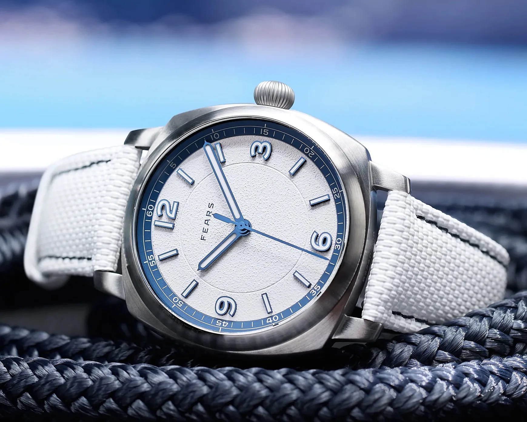 Close-up of a Fears wristwatch with a white textured dial, blue hour markers and hands, stainless steel case, and white fabric strap.