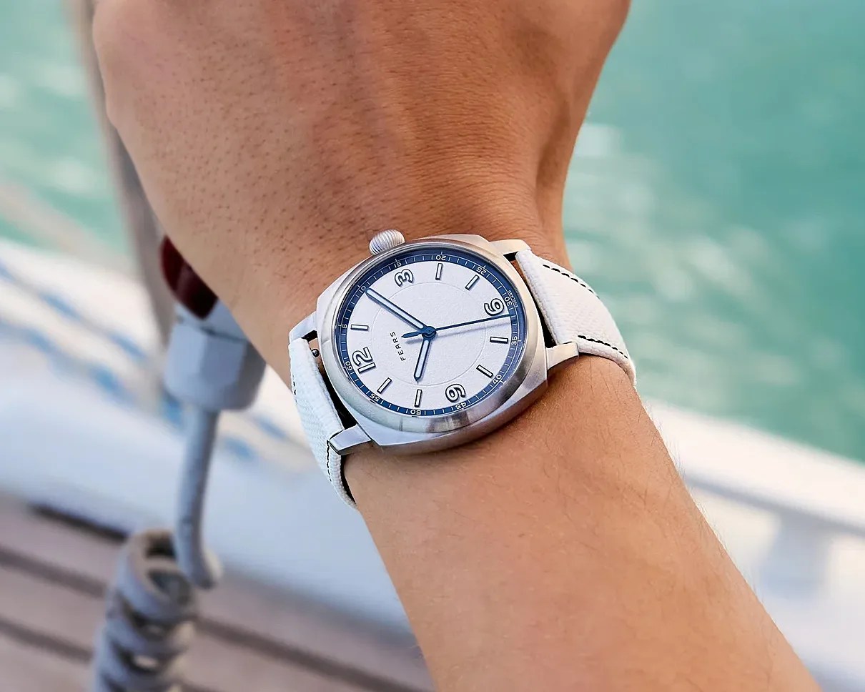 Silver wristwatch with white dial, blue accents, and white leather strap worn on a wrist near water.