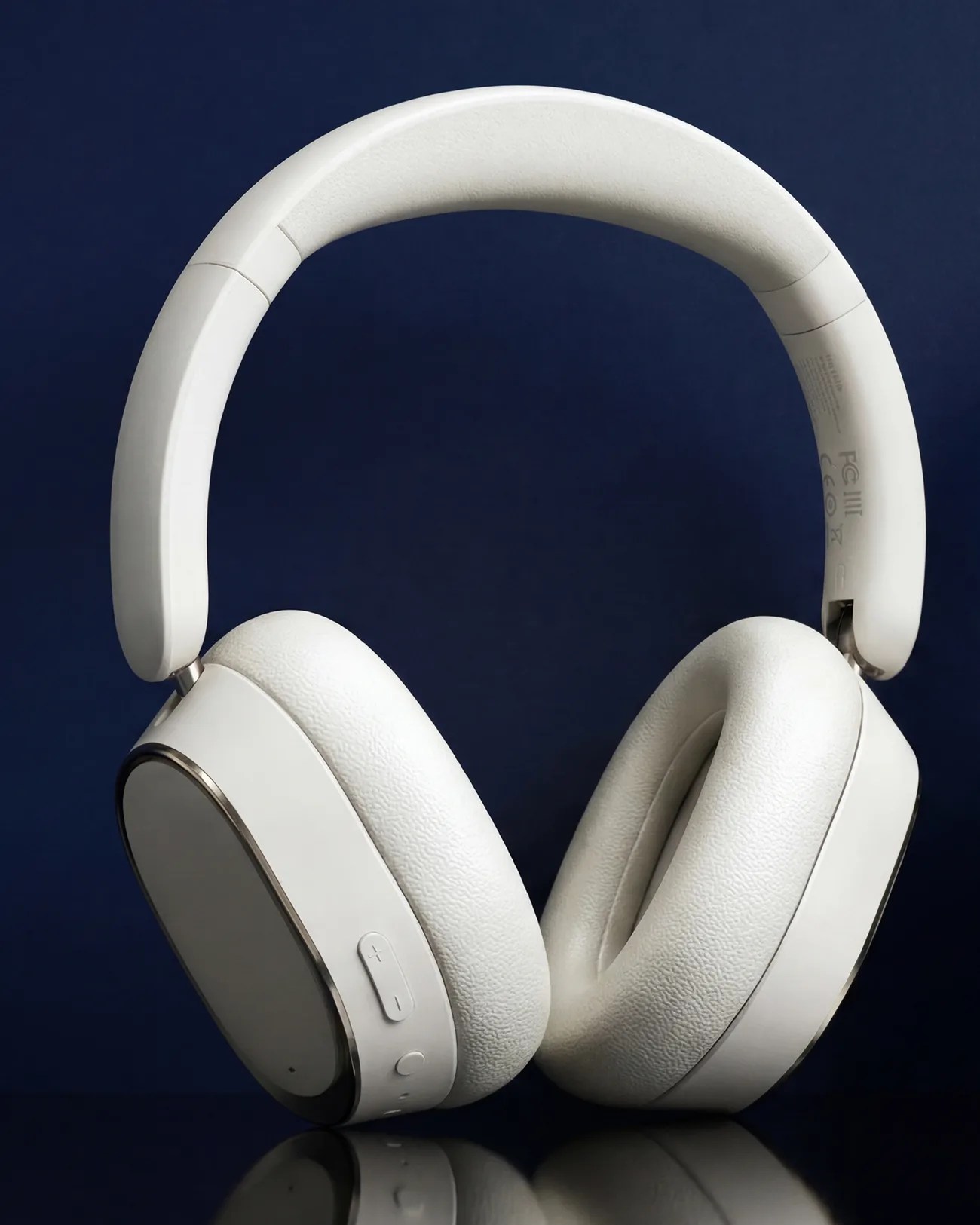 White over-ear wireless headphones with padded ear cushions and volume buttons on a dark blue background.