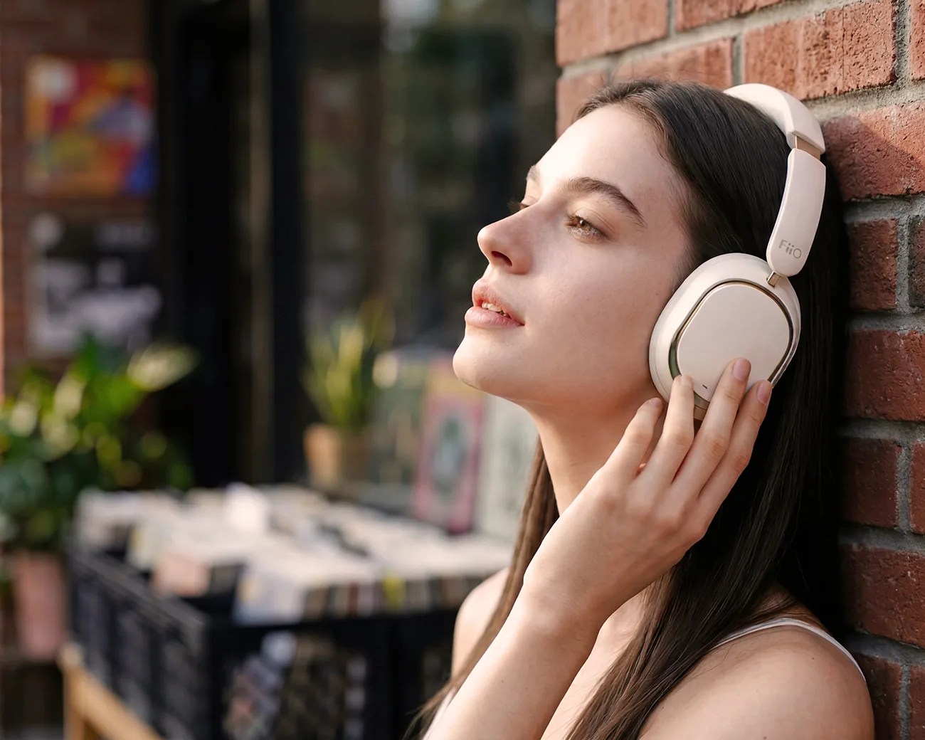 Person wearing white FiiO over-ear headphones, leaning against a brick wall with eyes closed.