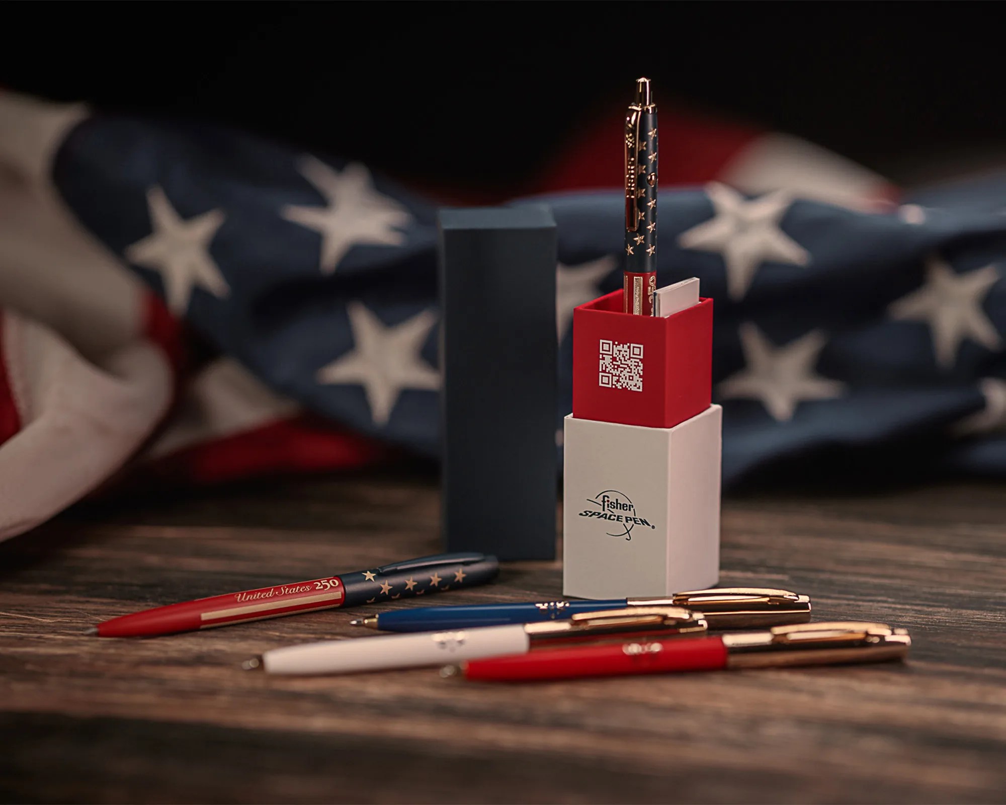Set of Fisher Space Pens with patriotic red, white, and blue colors and star designs, displayed on a wooden surface with an American flag in the background.