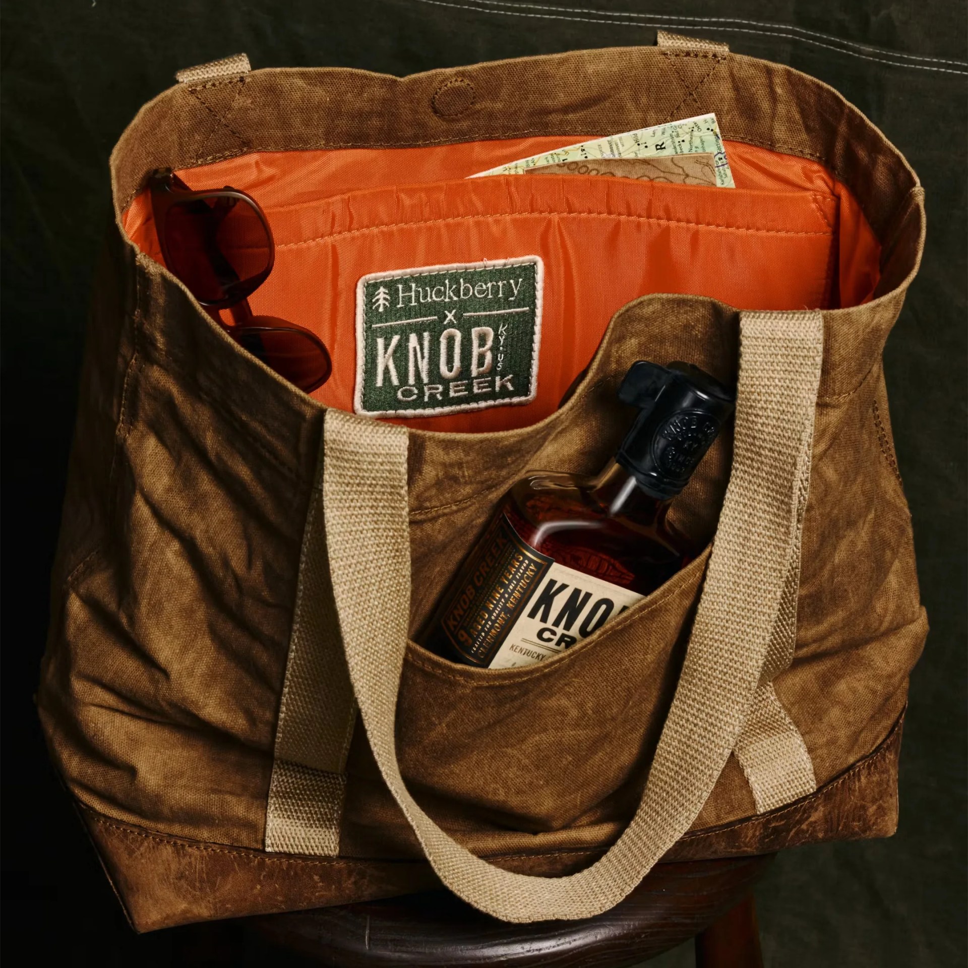 Brown canvas tote bag with beige straps, orange interior, sunglasses, a map, and a bottle of Knob Creek whiskey inside.