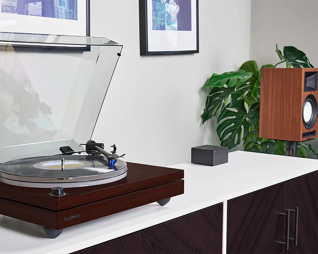 Wooden Fluance turntable with clear dust cover and black vinyl record on white cabinet next to wooden speaker and green plant.