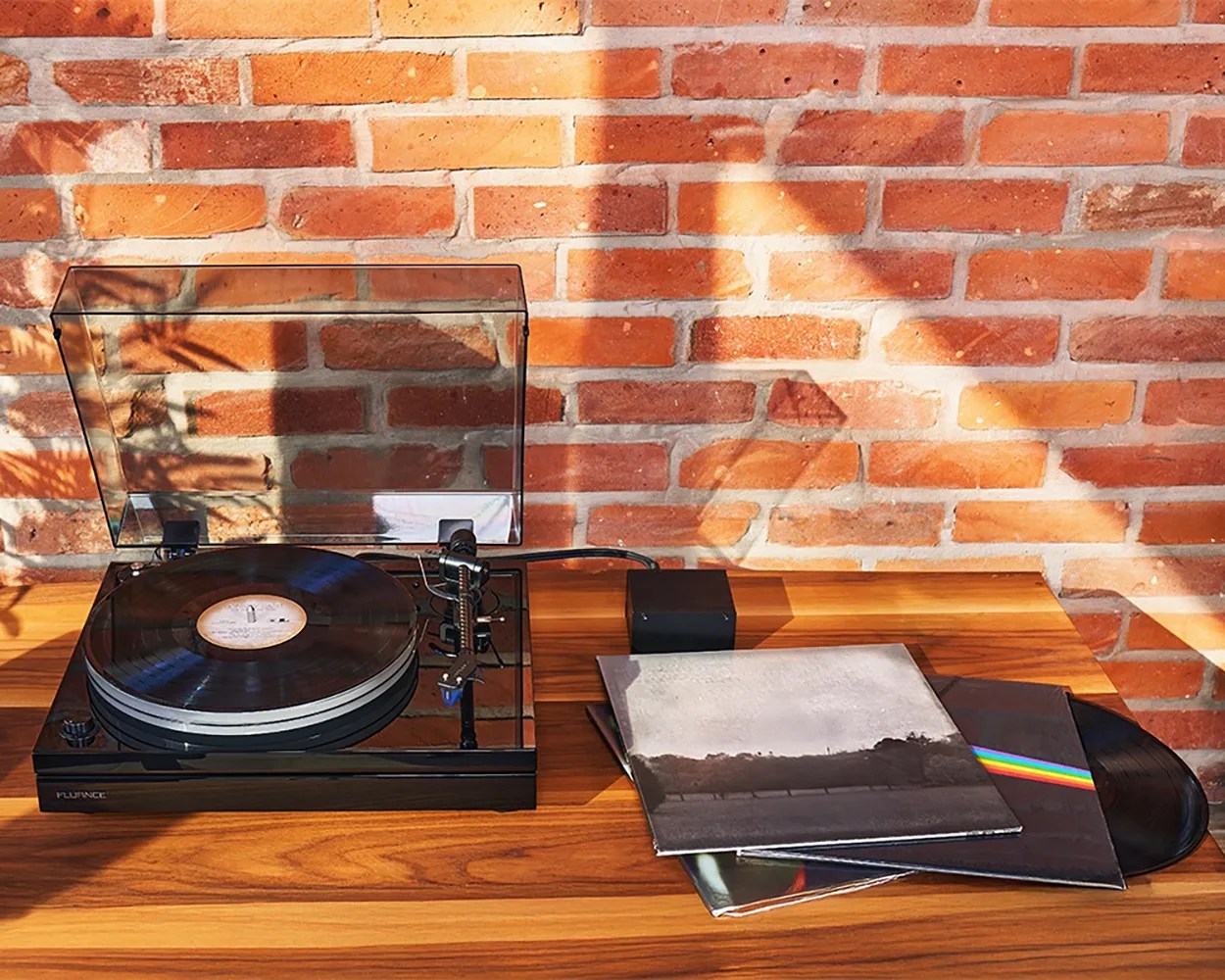 Black Fluance turntable with clear dust cover open, playing a vinyl record on a wooden surface next to three vinyl album covers.