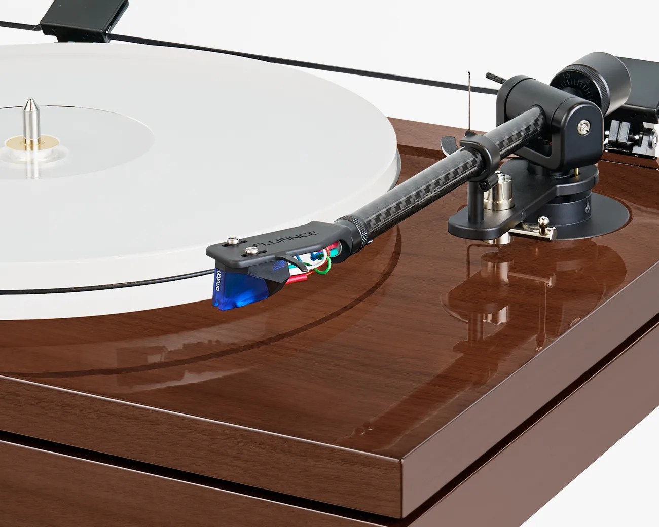 Close-up of a turntable tonearm with a blue Ortofon cartridge on a polished wood base and a white vinyl record.