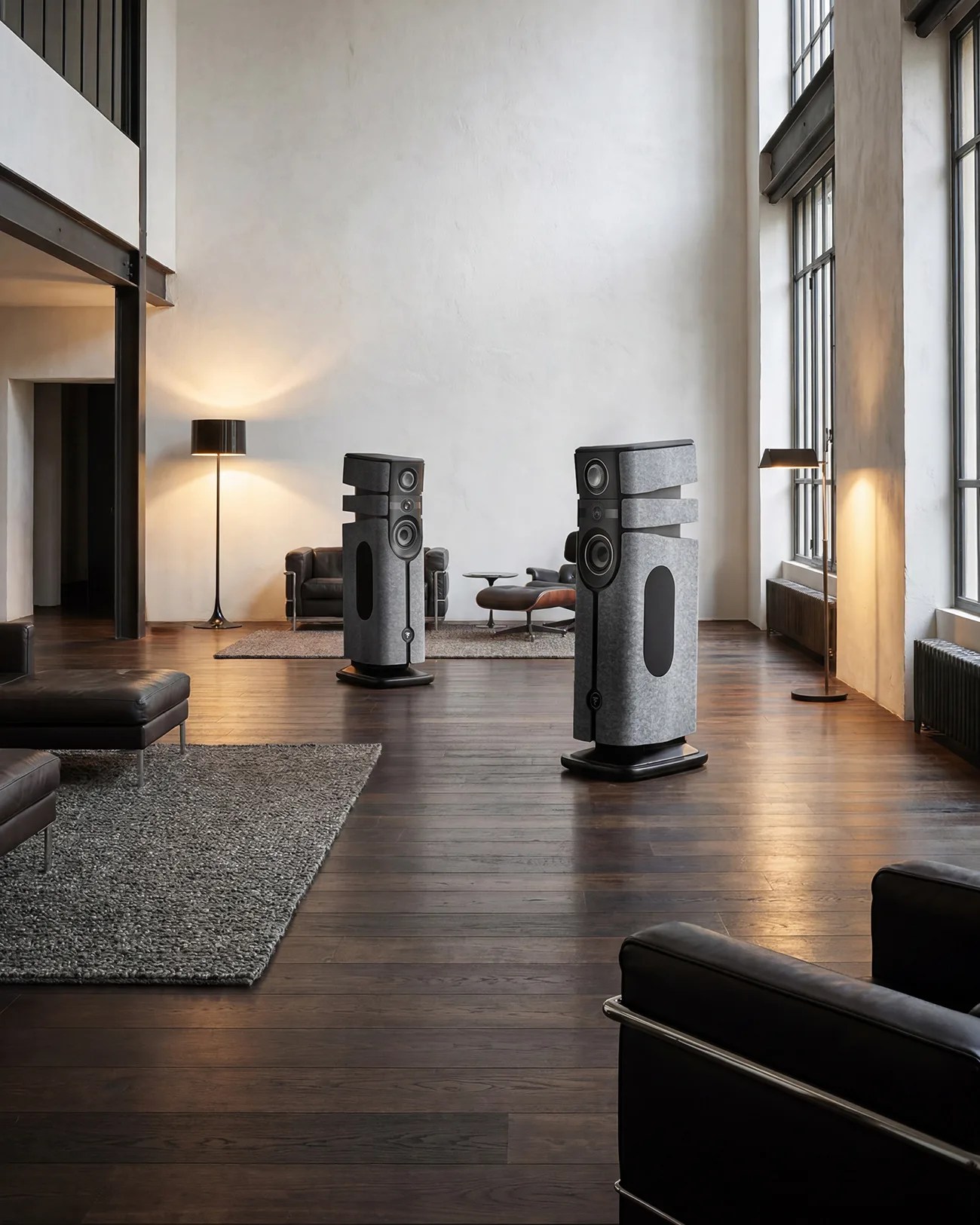 Pair of large gray floor-standing speakers with black accents in a modern living room with wooden floors and leather furniture.