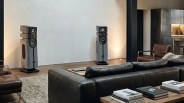 Pair of large gray floor-standing speakers with black accents in a modern living room with leather sofas and wooden furniture.