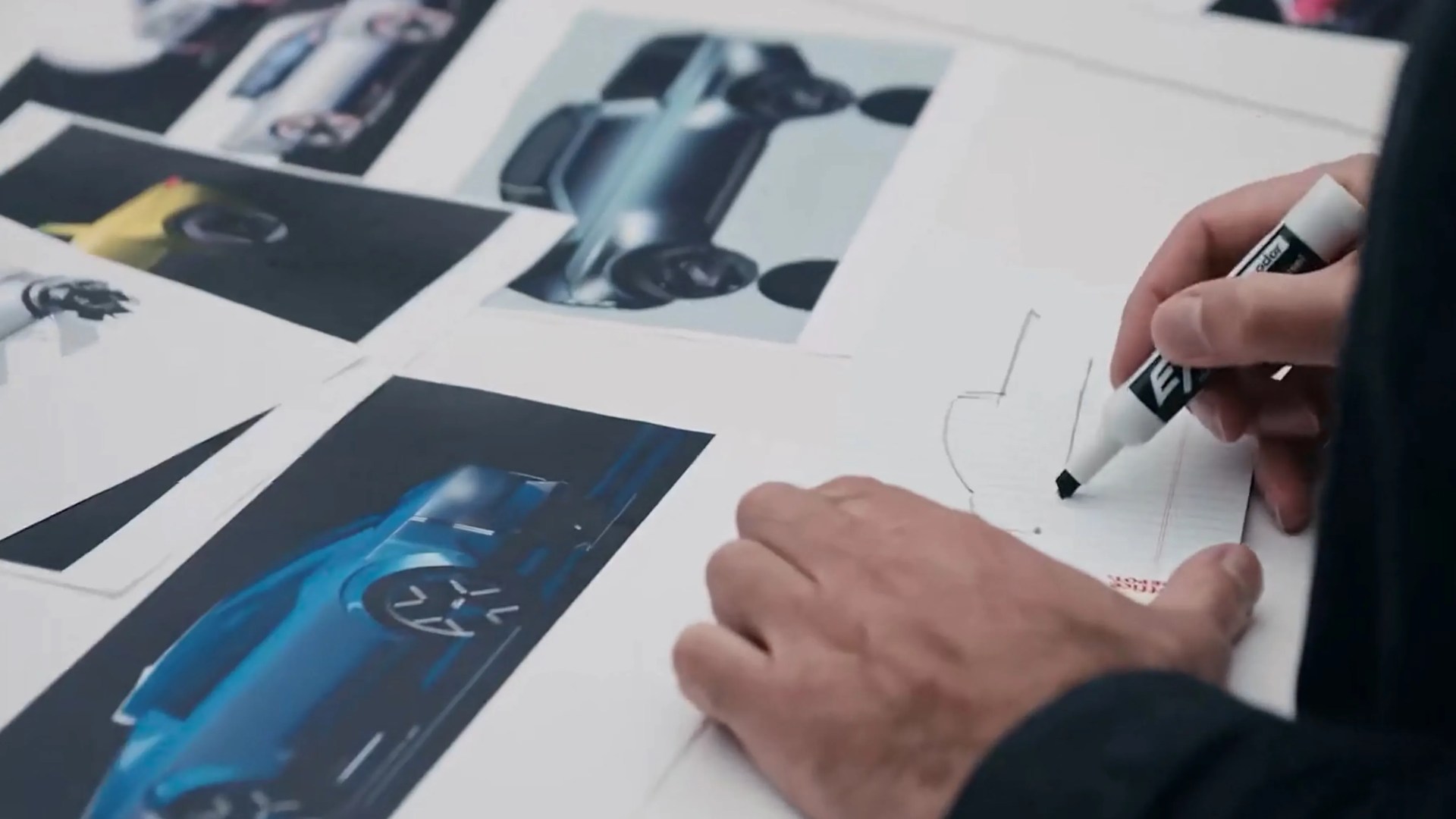 Hands sketching a car design on paper surrounded by printed car concept images.