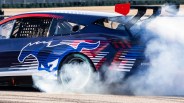 Blue and red Ford Mustang race car drifting with smoke around rear tire and large rear spoiler.