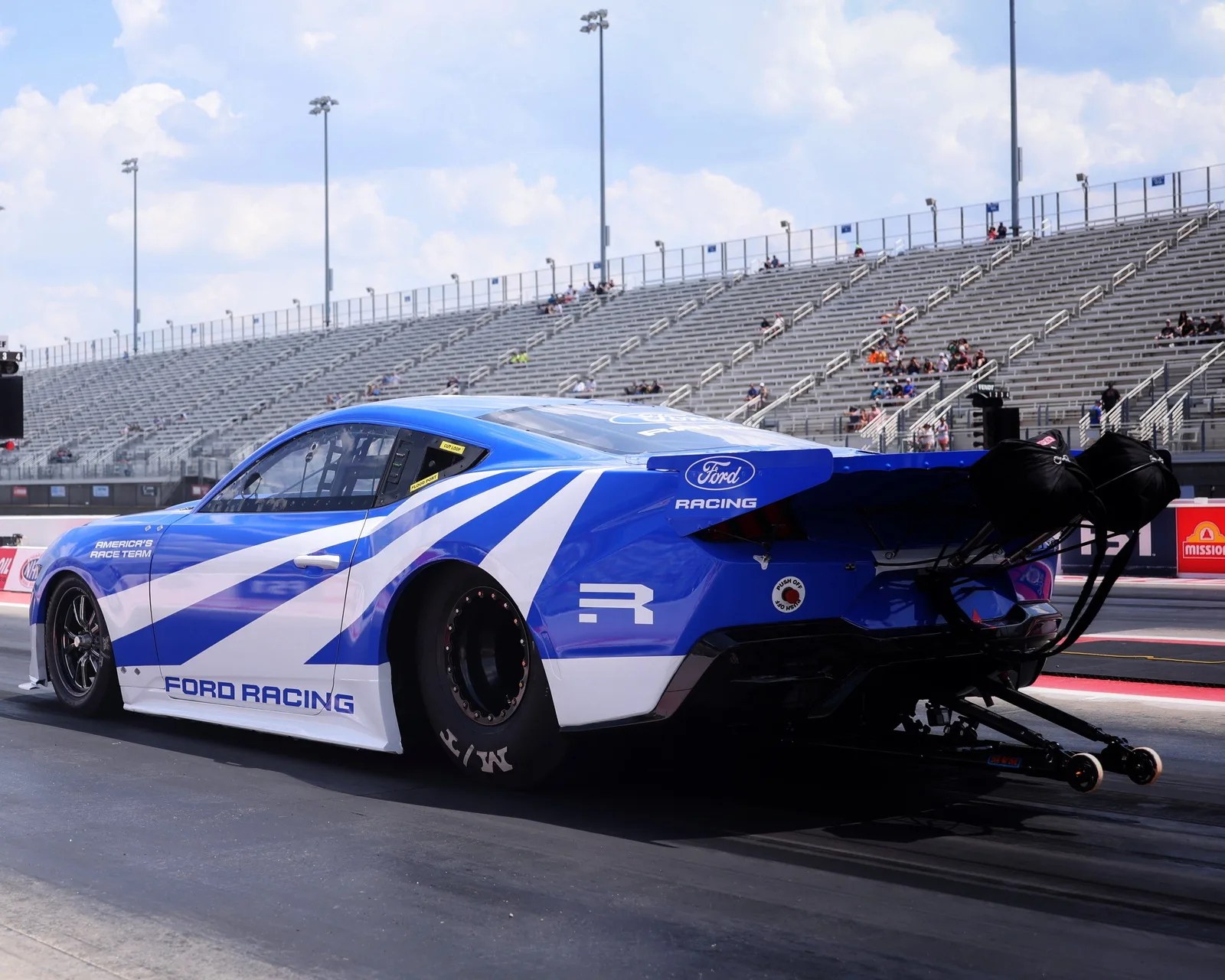 Blue and white Ford Racing drag race car with parachutes on a racetrack with empty bleachers in the background.