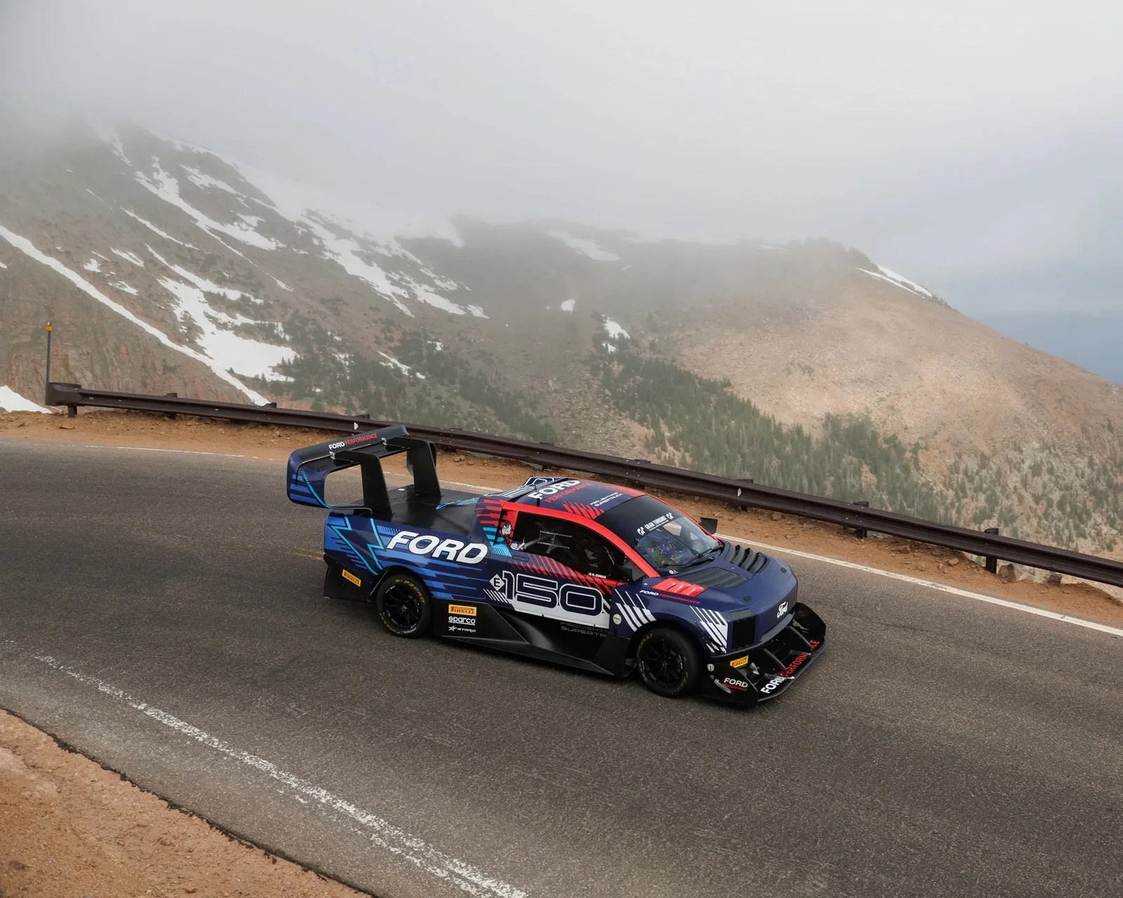 Blue and red Ford race truck with large rear wing on a mountain road with guardrails and foggy peaks.